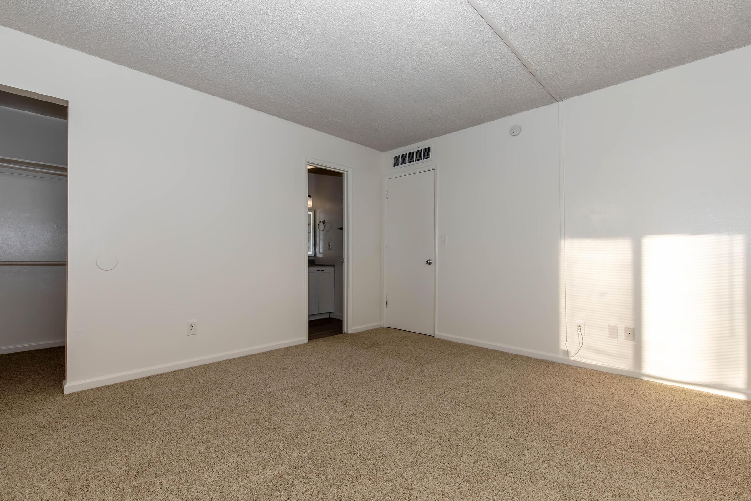 A spacious, empty room with light-colored walls and beige carpet. A door leads to a bathroom or another room, and another door is visible on the left, suggesting a closet. Natural light streams in through a window, casting a soft shadow on the wall.