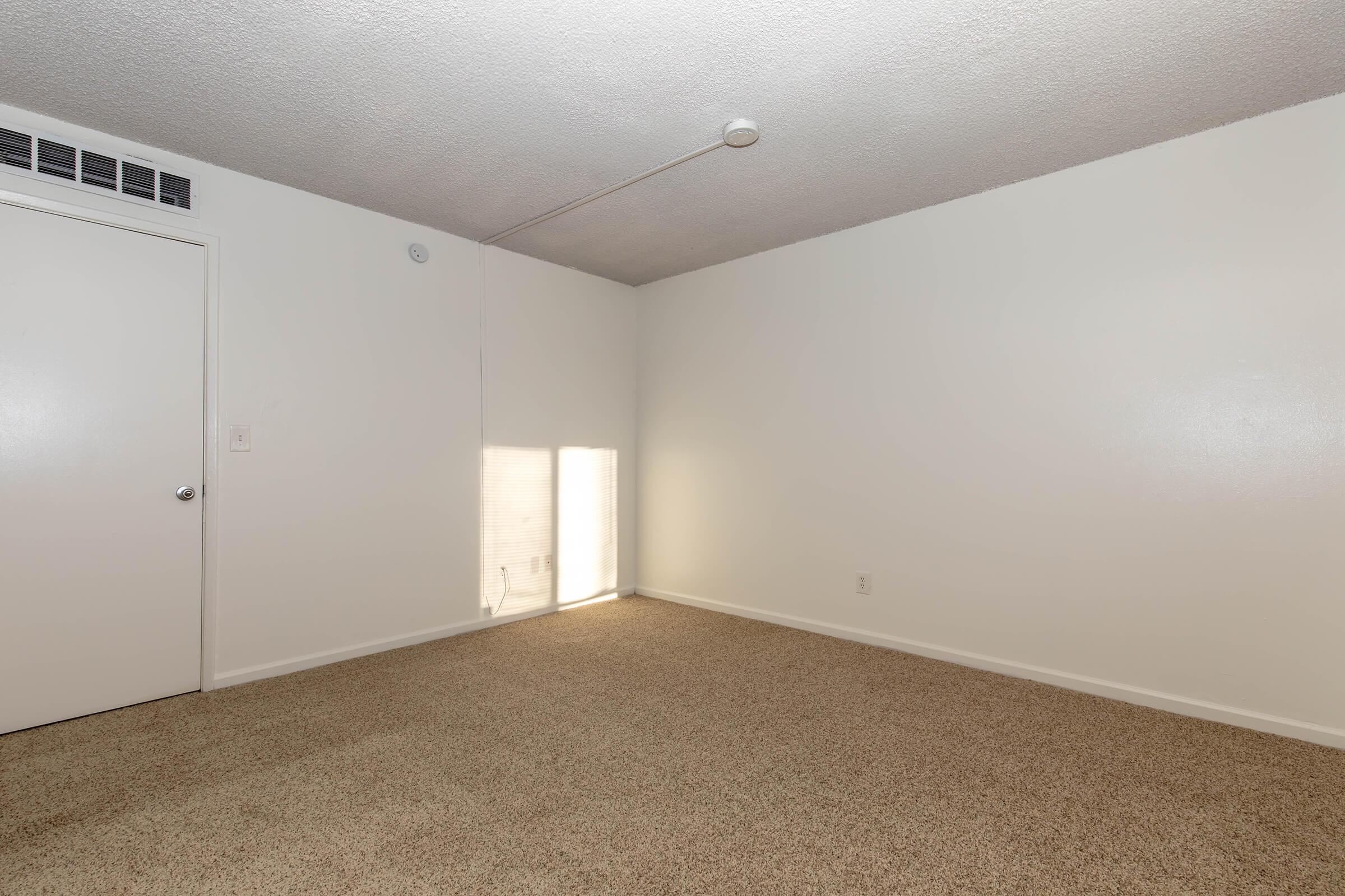 A sparsely furnished room with a light-colored carpet and white walls. There is a closed door on the left and a small window on the right, allowing natural light to enter. The ceiling appears textured, and a single ceiling light fixture is visible. The overall ambiance is bright and airy, suggesting a clean, neutral space.