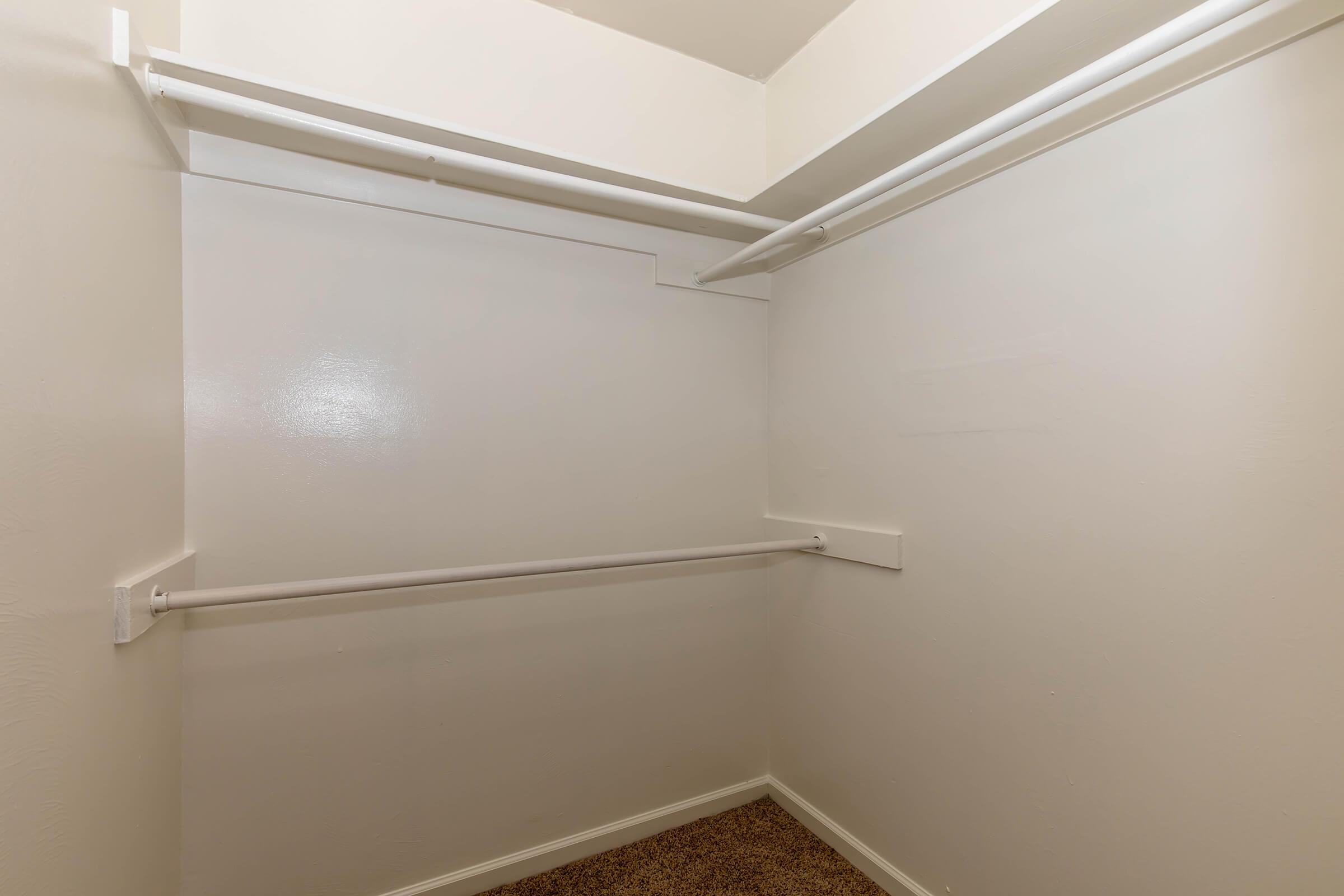 Empty closet with two horizontal hanging rods against plain white walls and a carpeted floor, providing a simple storage space.