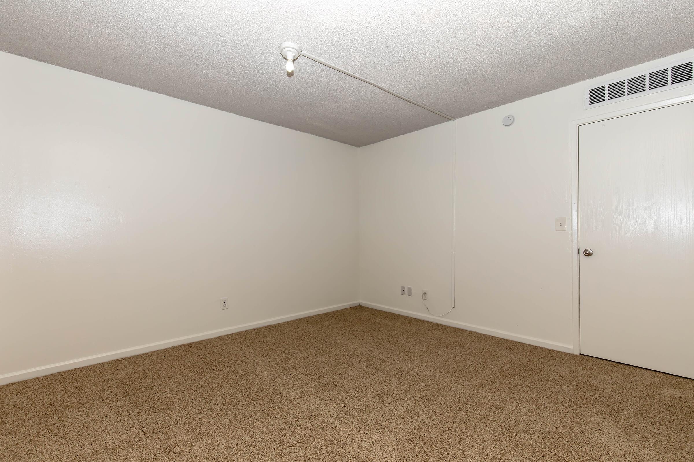 Empty room with beige carpeting and plain white walls. A single overhead light fixture is visible, and there's a closed door on the right side of the image. The space appears well-lit and clean, with no furniture or decorations present.