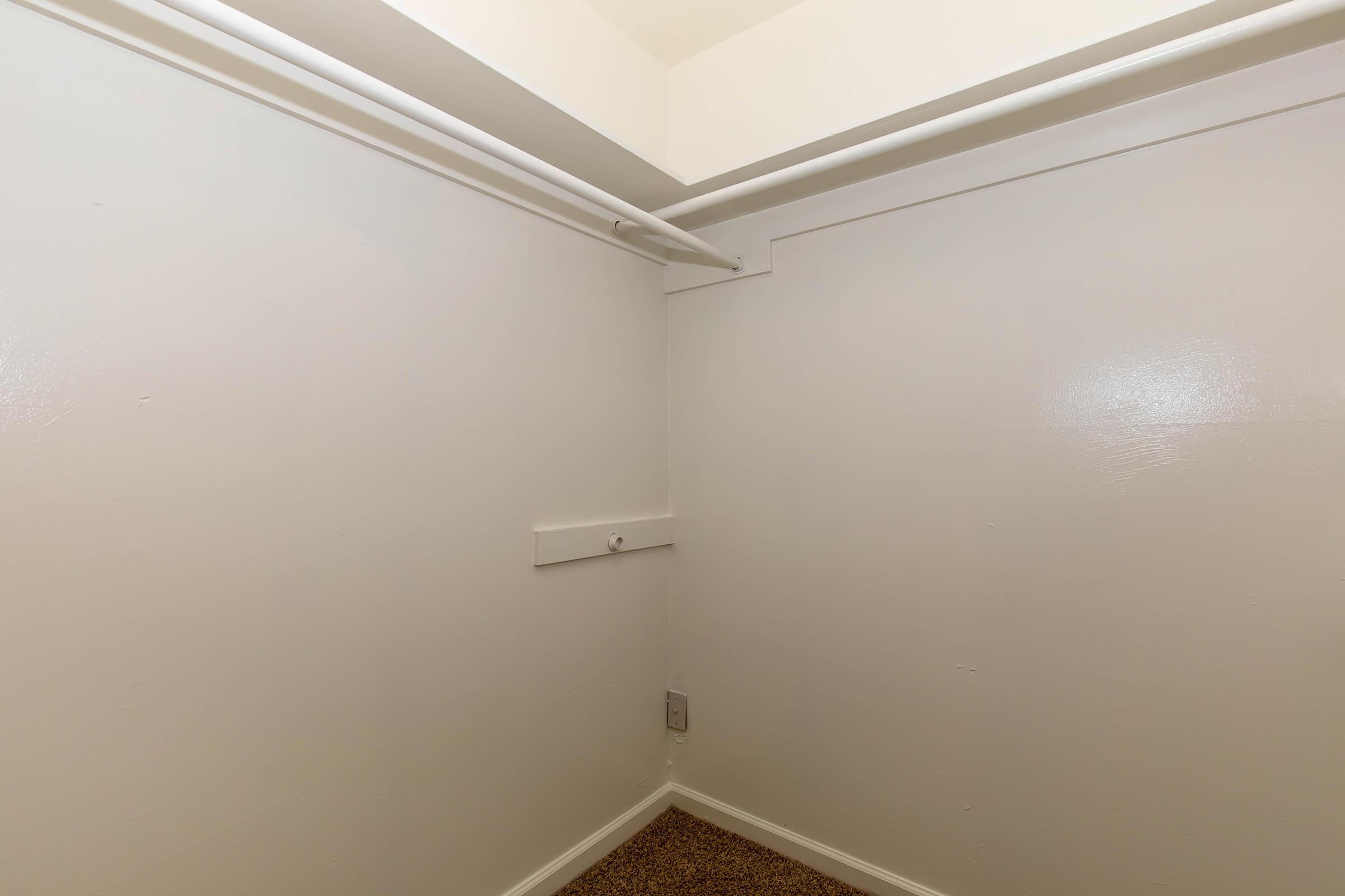 An empty corner of a closet featuring two white wall sections and a clothing rod mounted near the ceiling. The floor is covered with a brown carpet, and the walls are painted white, creating a clean and uncluttered appearance.