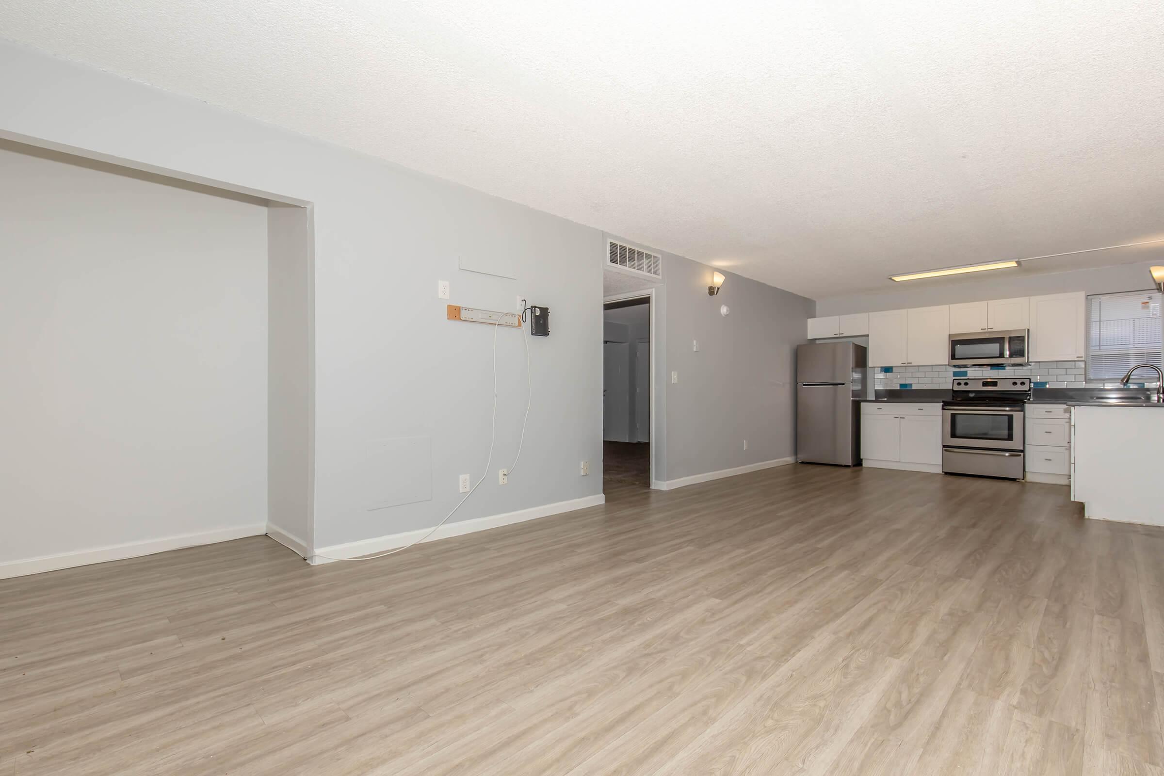 Spacious open-concept living area with light gray walls and wooden flooring. The kitchen features modern stainless steel appliances, including a refrigerator and oven. Ample natural light fills the space, enhancing the airy atmosphere. Wall-mounted cable provider box visible on the left side.