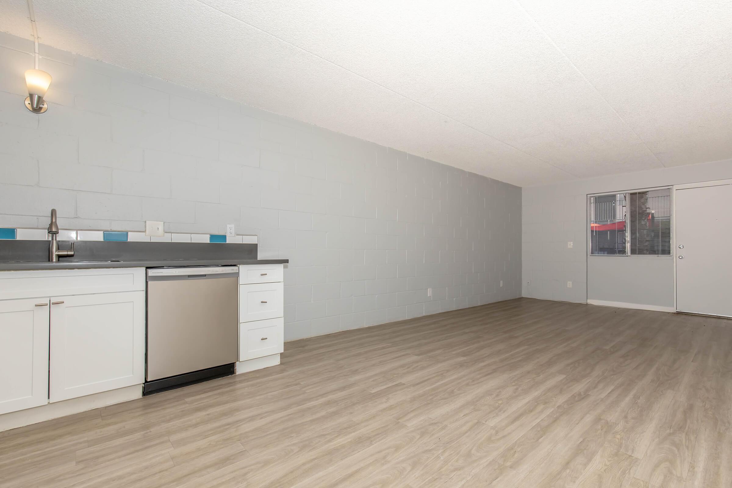 A spacious, empty room featuring a kitchen area along one wall with a stainless steel dishwasher, white cabinetry, and a gray countertop. The floor is light-colored wood laminate, and there's a large window allowing natural light, with a door leading outside. The walls are painted light gray.