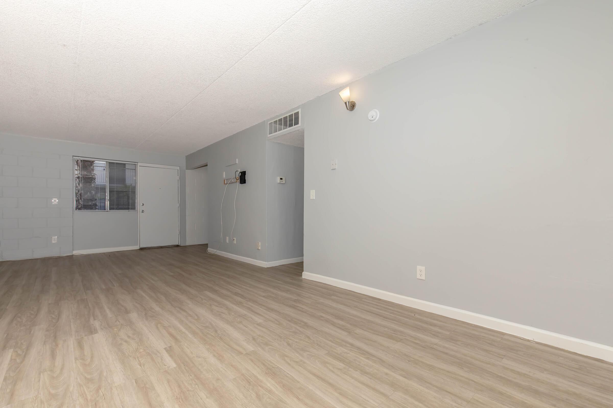 A spacious, empty room with light gray walls and laminate flooring. A door leads to an entrance, with a small wall-mounted device nearby. Natural light enters through a window, illuminating the space, which has a simple, clean aesthetic without furnishings.