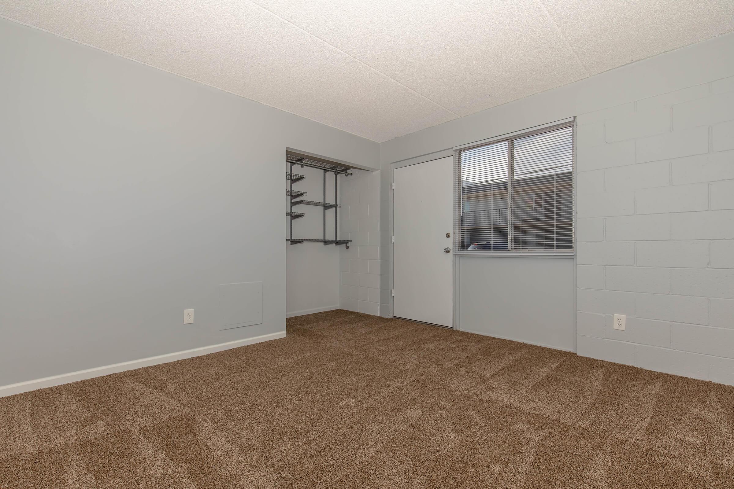 A well-lit, empty room with light gray walls and carpet. There is a closet with shelving on one wall and a door leading outside. A window allows natural light to enter, enhancing the spacious feel of the room, which has a clean and modern appearance.