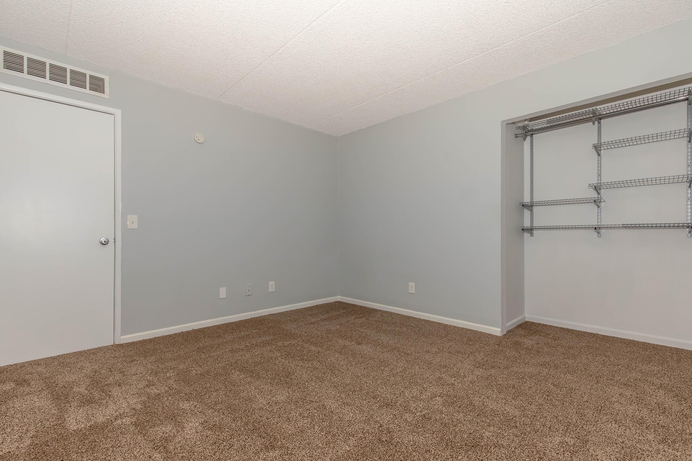 Empty room with light gray walls, carpeted floor in a neutral tone, and a small closet with metal shelving. A door is visible on the left. The space appears clean and ready for furniture or decoration. Natural light may come from a window not depicted in the image.