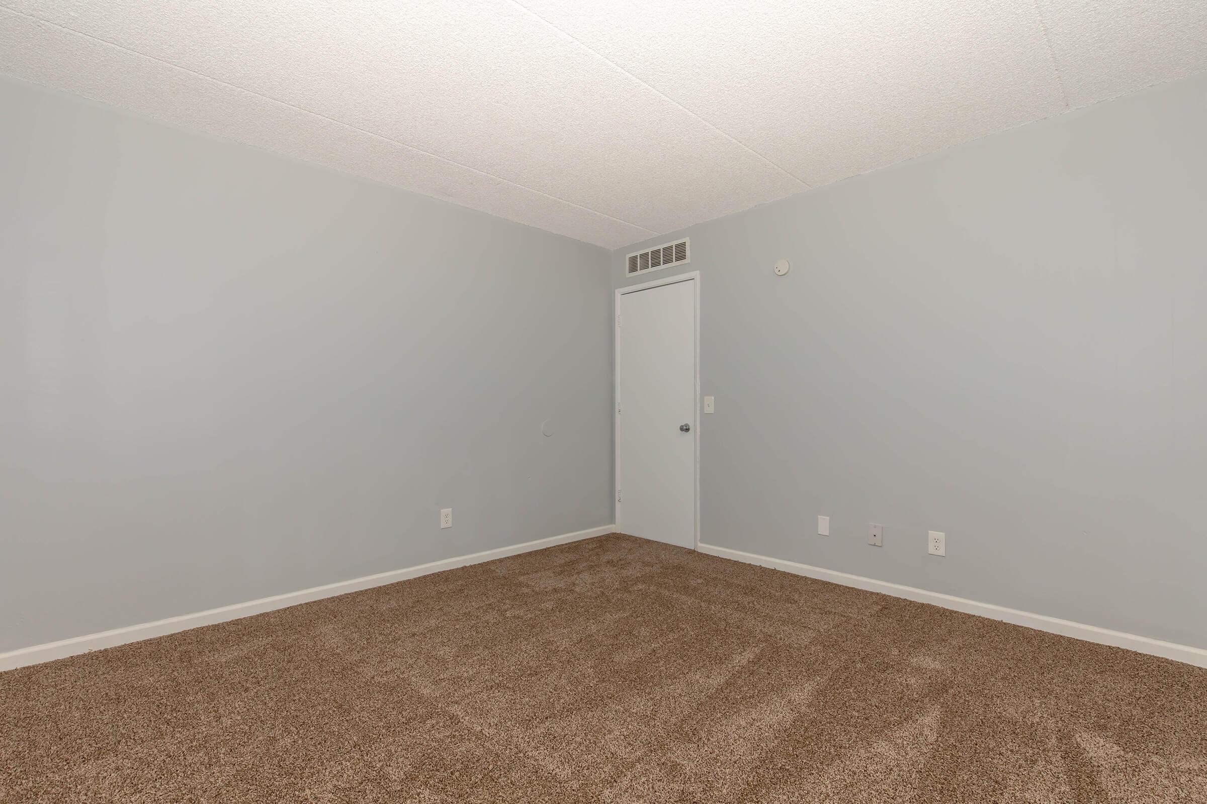 Empty room with light gray walls and beige carpet. The space features a single door on one wall and a small vent near the ceiling. The lighting is soft, creating a neutral and inviting atmosphere. No furniture or decor is present, emphasizing the room's simplicity and potential for personalization.