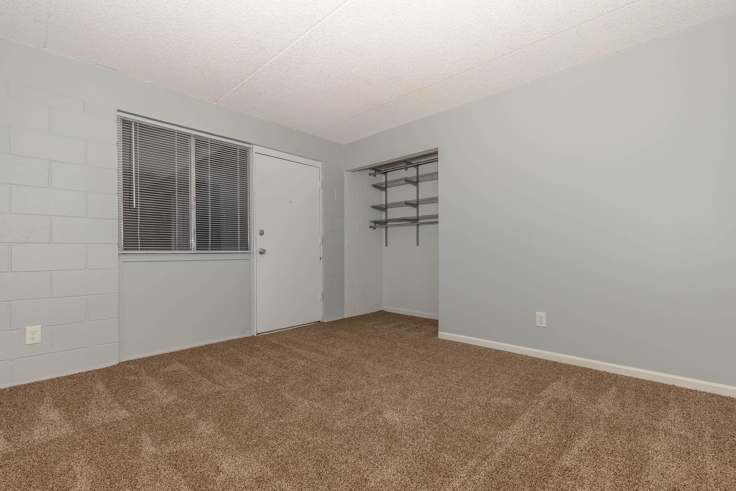 Empty room with gray walls and a light gray ceiling. The floor is covered in brown carpet. A small window with vertical blinds is on one side, and a door leads outside. There are shelves along one wall, with an alcove next to the door. The overall atmosphere is minimalist and clean.
