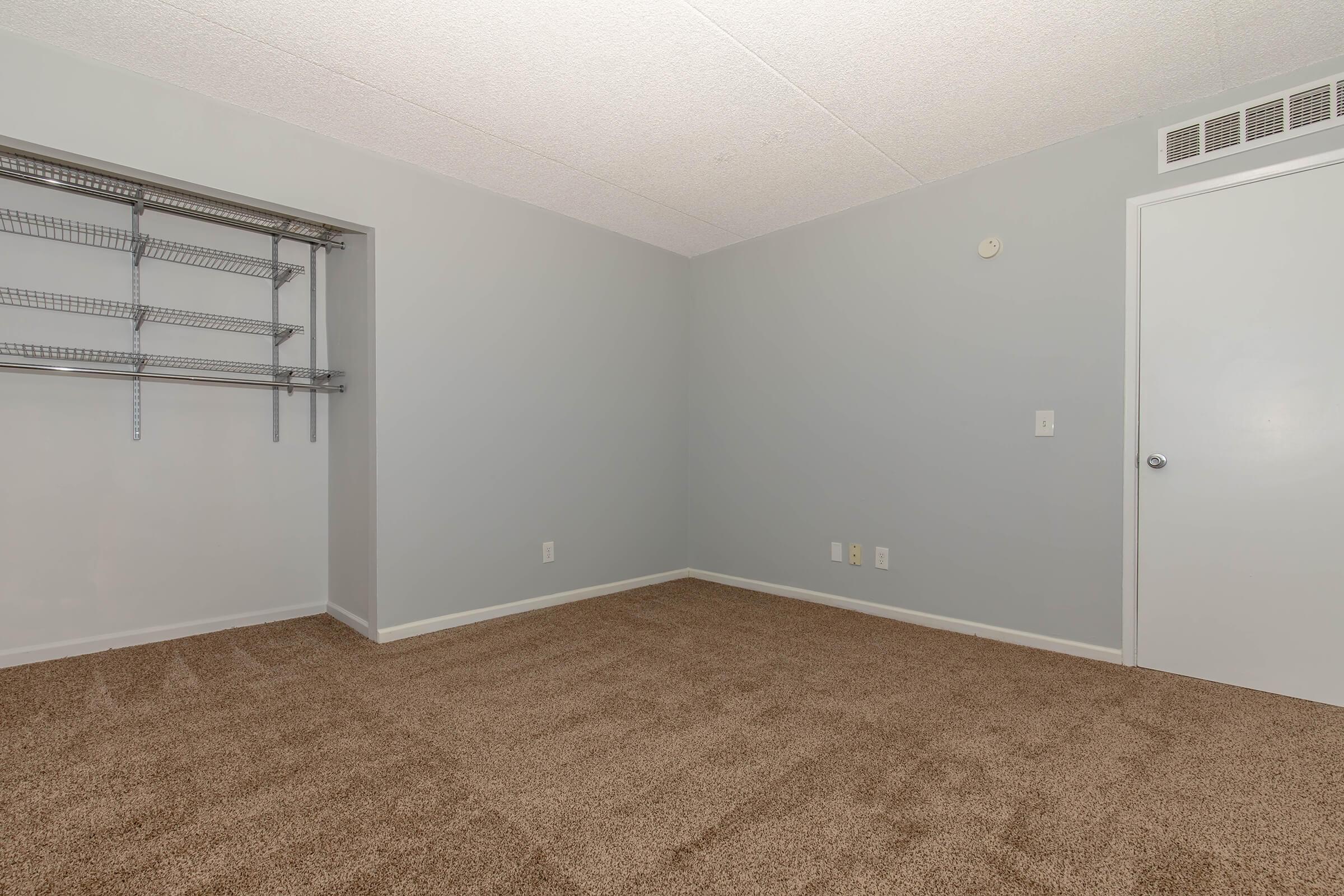 Empty room featuring light gray walls and a beige carpet. There is a metal shelving unit mounted on the wall to the left, and a closed door on the right. The room is well-lit with no furniture, creating a spacious and minimalist atmosphere.