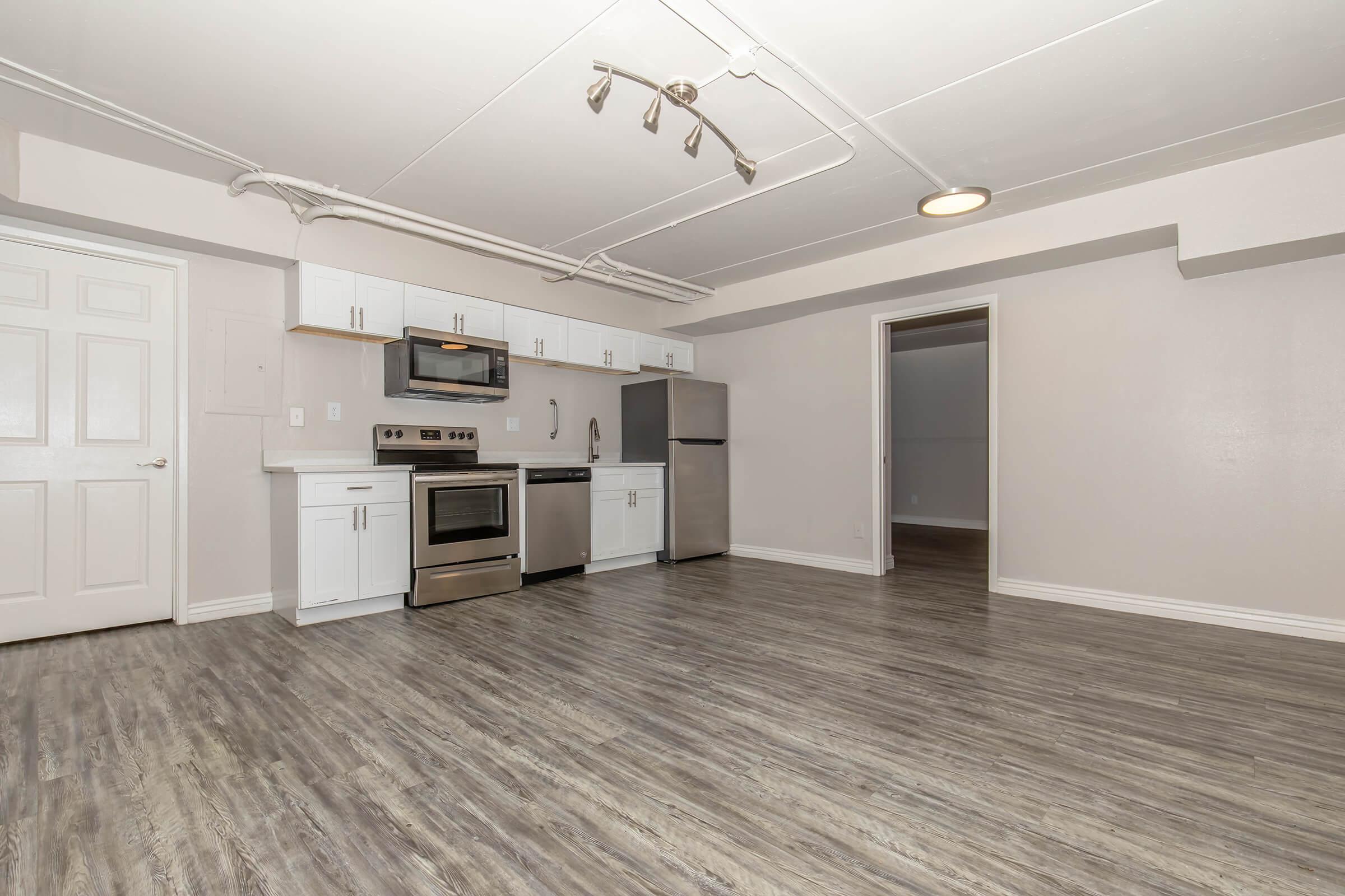 A modern kitchen in a bright, open space featuring stainless steel appliances, white cabinetry, and light-colored walls. The floor is a wood-like laminate, and there is track lighting overhead. A door leads to another room, and the layout creates a spacious feel.