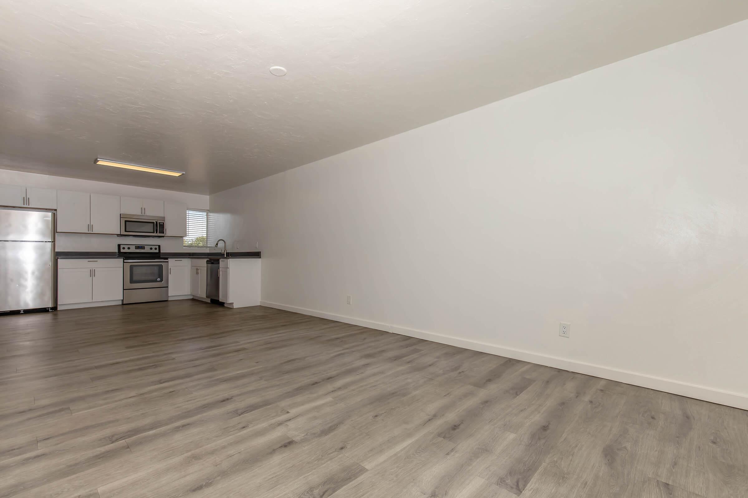 Spacious interior of a modern kitchen and living area, featuring light-colored walls, new cabinetry, stainless steel appliances, and a wood-like floor. Abundant natural light enters through a window, creating an inviting atmosphere.