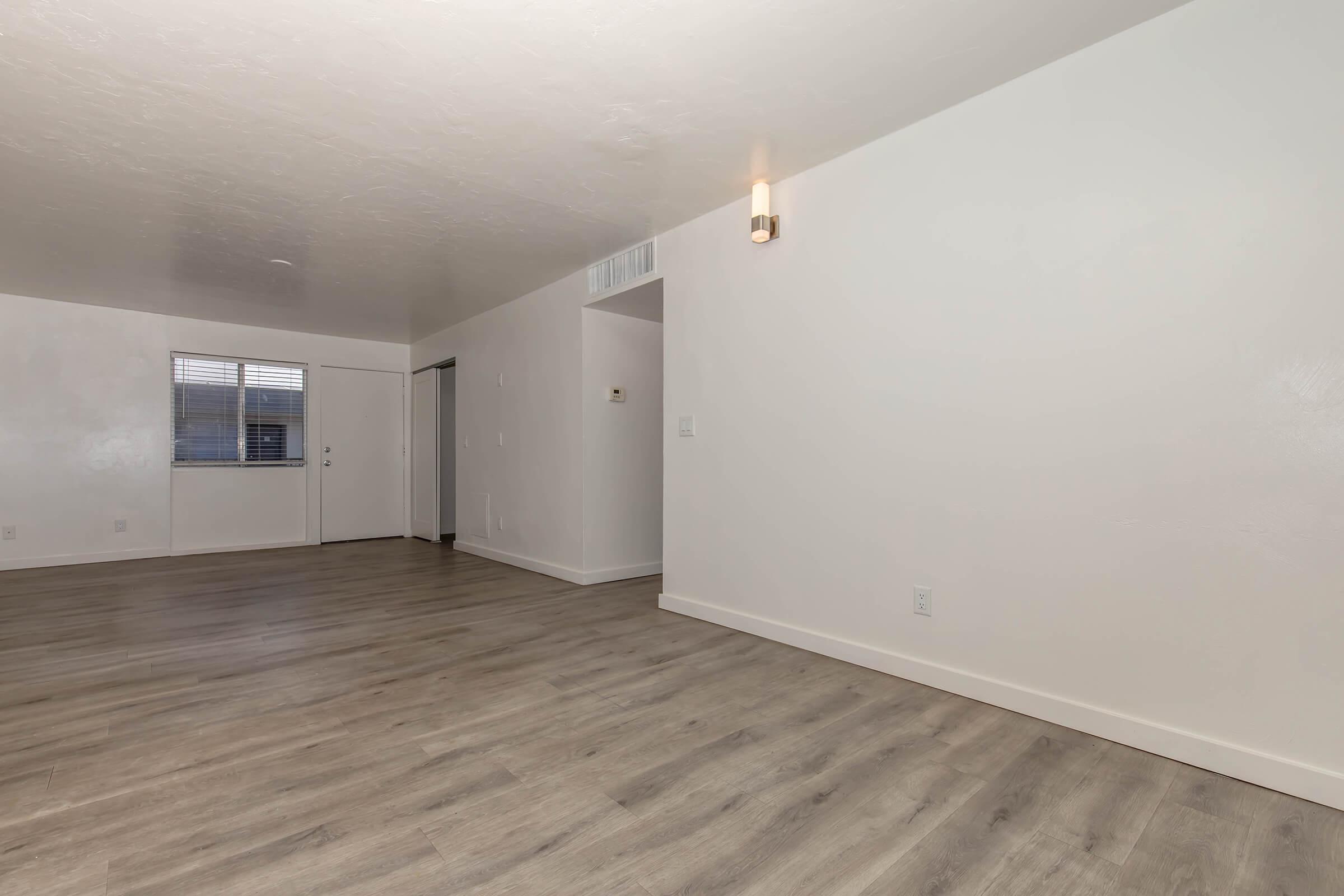A spacious, empty room with light-colored walls and brown laminate flooring. A small window allows natural light, and a door leads outside. One wall features a modern light fixture. The space is suitable for various furniture arrangements and decor.