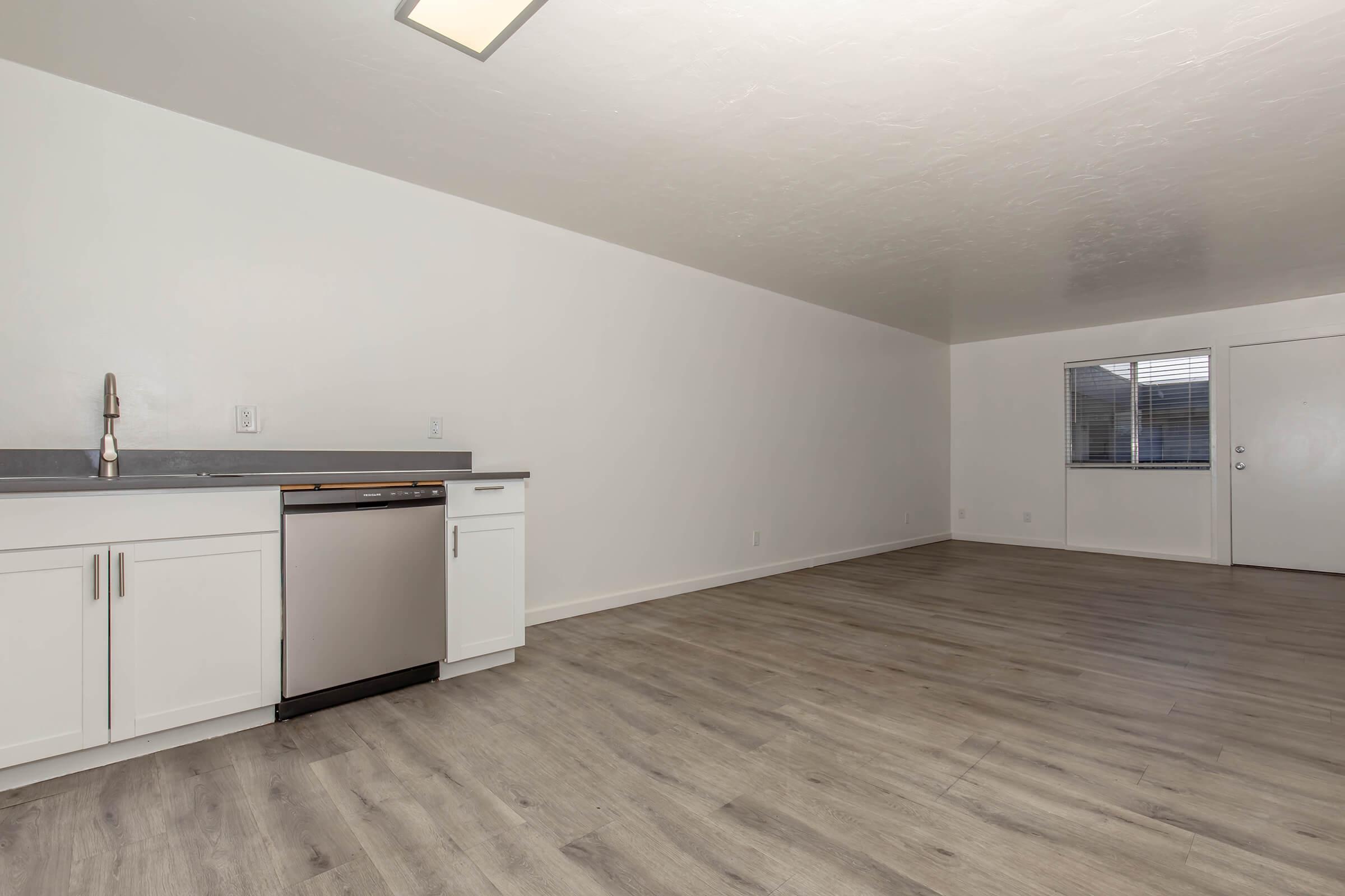 A spacious, minimally furnished room featuring a modern kitchen area with a stainless steel dishwasher. The walls are painted white, and the floor has a light wood-like finish. A window allows natural light to enter, and a door leads outside. The space is uncluttered, ideal for various design possibilities.