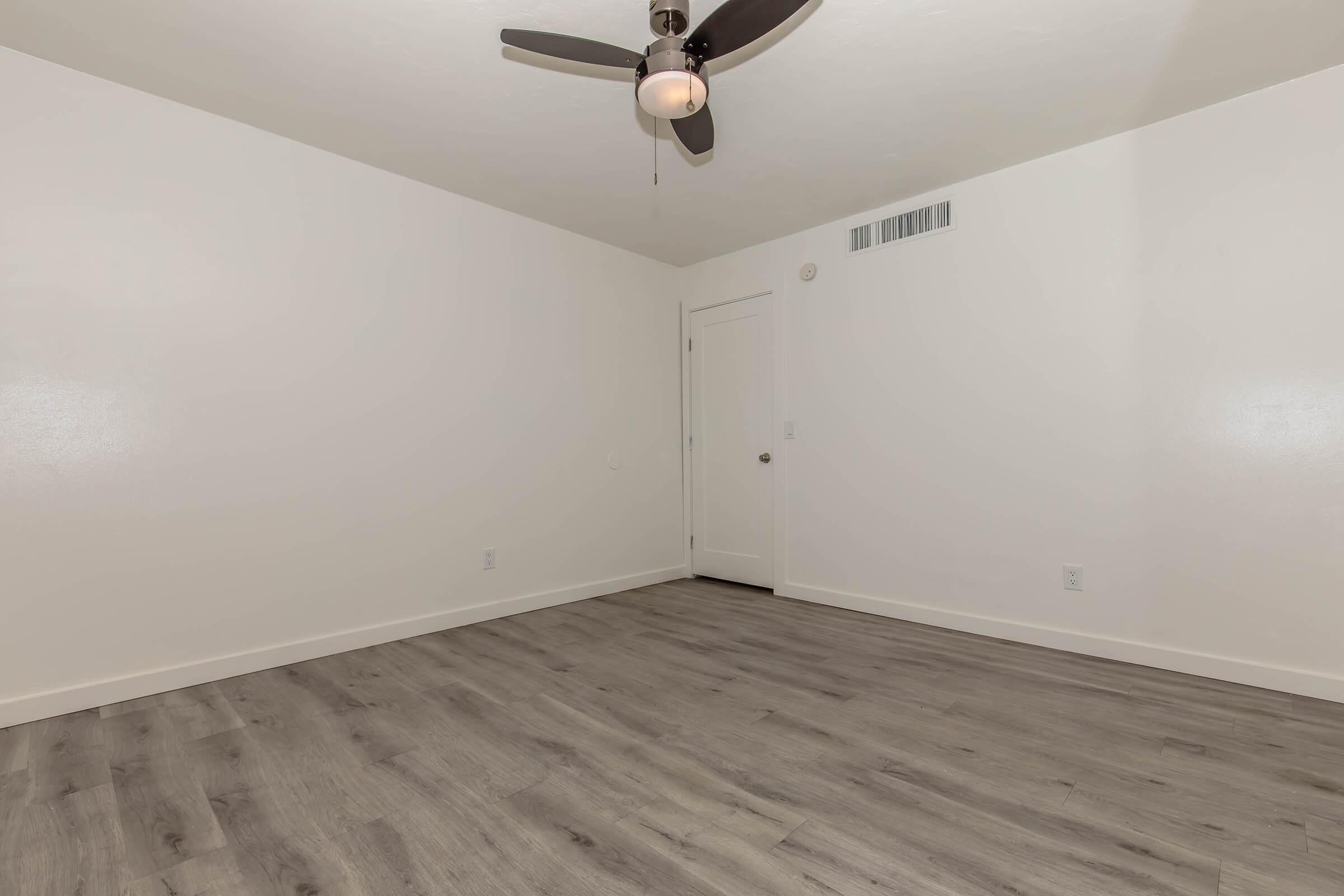 Empty room with light-colored walls and a ceiling fan. There is a plain white door on the far wall, and the floor is covered in laminate that mimics wood. The space is well-lit and features a minimalist design.