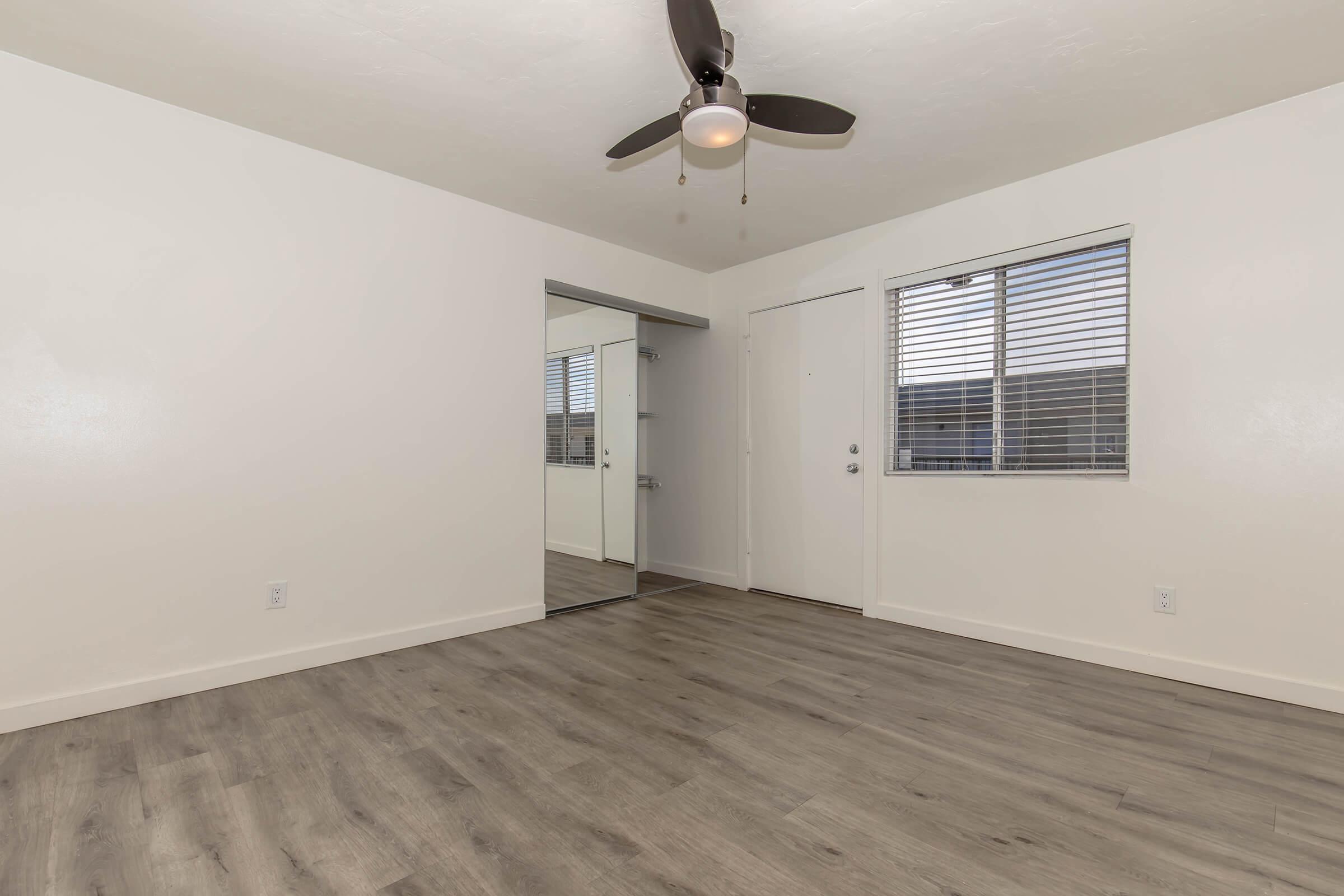 A spacious, empty room with light-colored walls and a wooden floor. It features a ceiling fan with a light, a large mirror on one wall, and a window with horizontal blinds. The door to the room is visible, alongside natural light coming in from the window.