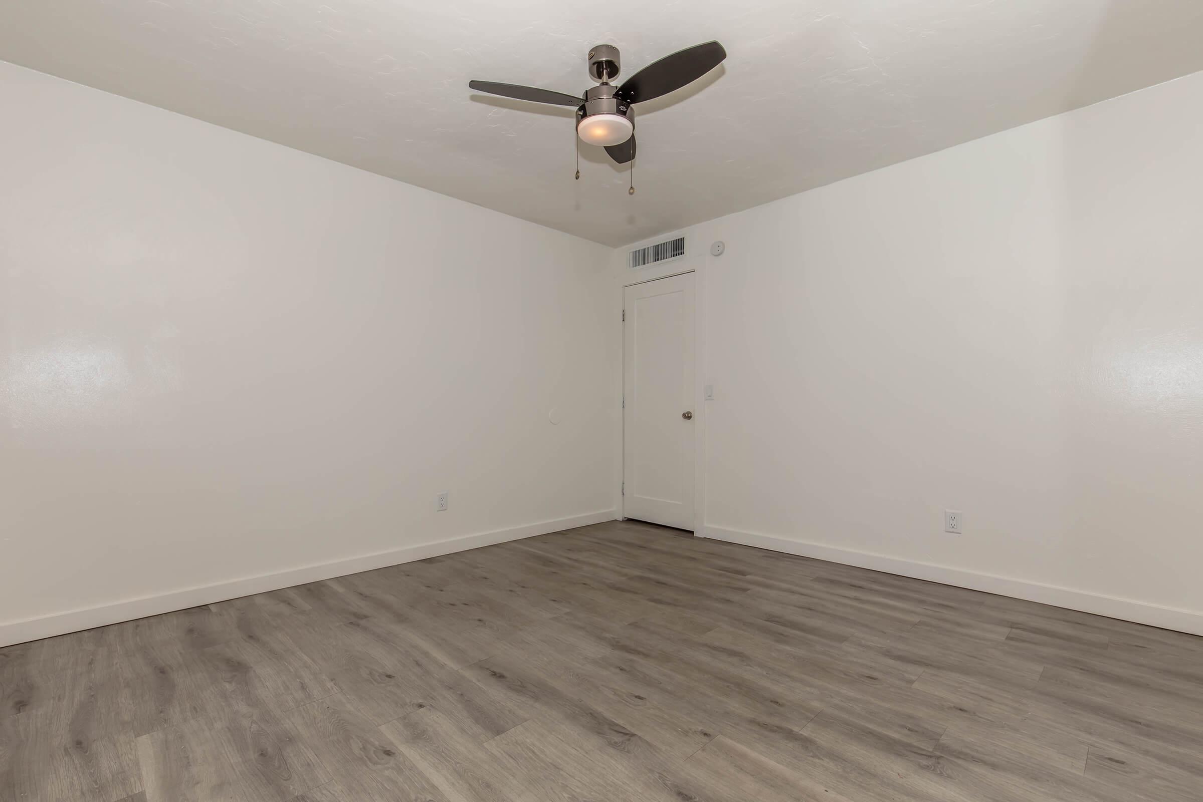 Empty room with light-colored walls and a light brown wood-like floor. A ceiling fan with three blades hangs from the ceiling. There is a white door on the right side, leading to another space. Natural light illuminates the room, creating a bright and open atmosphere.