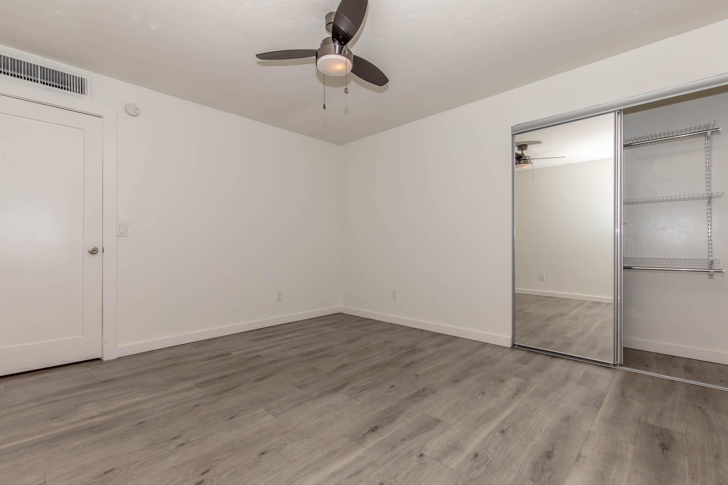 A bright, empty room with light-colored walls and a ceiling fan. The floor is covered with light wood-like laminate. One wall features a sliding mirrored closet door, while another wall has a plain door leading to another area. The space appears clean and ready for furnishing.