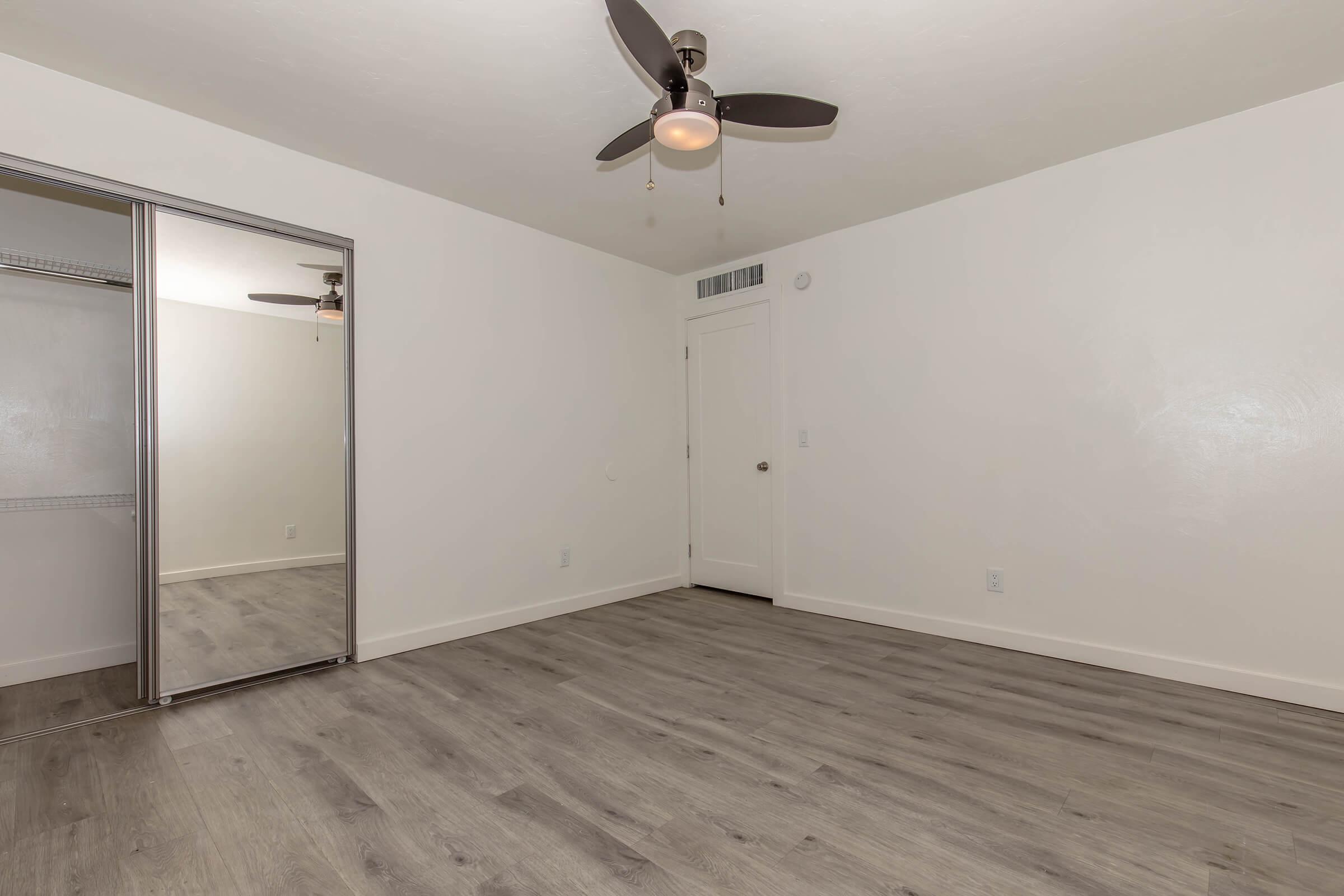 A spacious, empty bedroom featuring light-colored walls and a ceiling fan. There is a mirror closet on one side and a door leading out of the room. The flooring is light wood, enhancing the bright and airy atmosphere of the space.