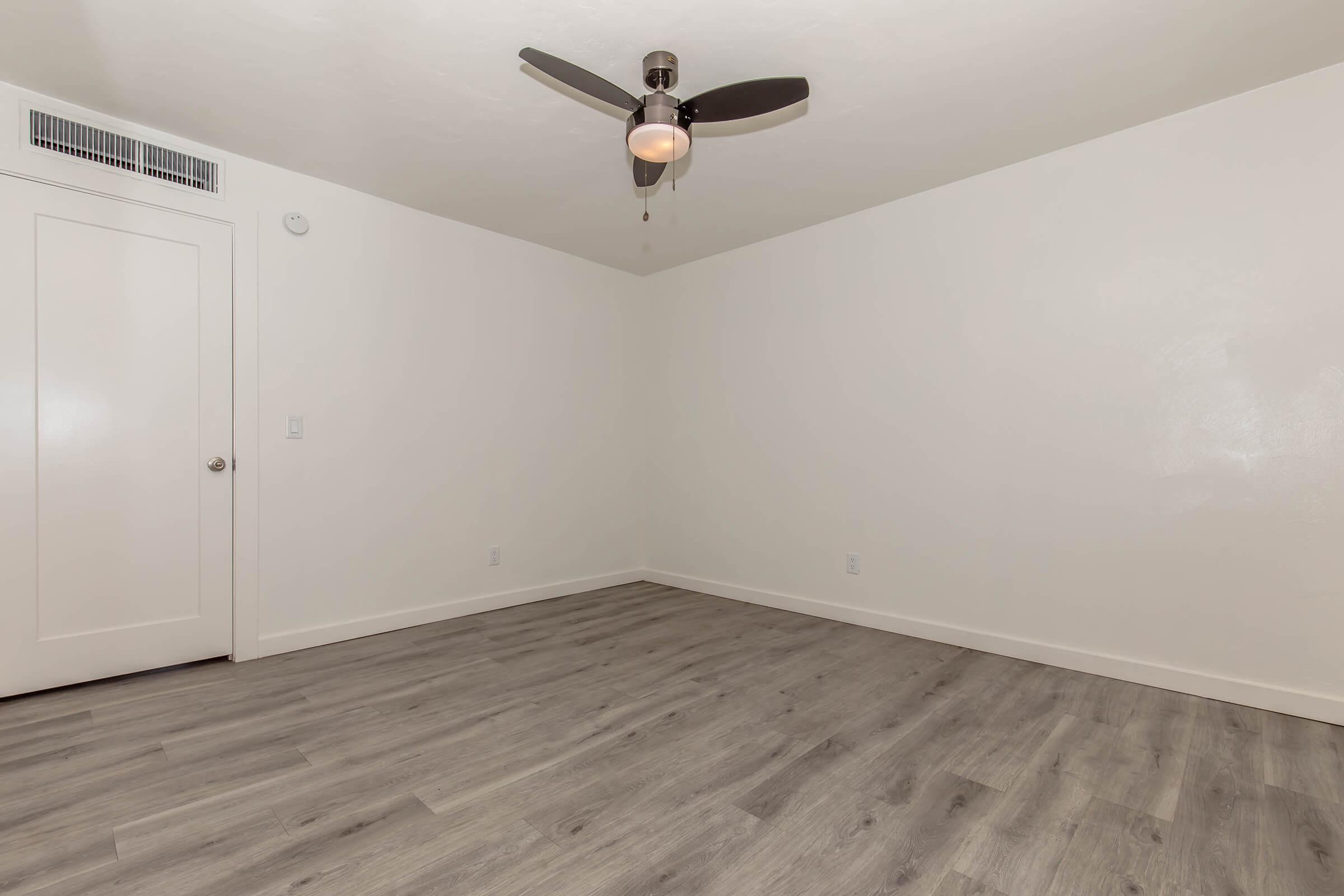 A spacious, empty room with light-colored walls and a light wood floor. Features a ceiling fan with a light fixture and a white closet door on the left. The room has a bright and airy feel, ideal for personalization.