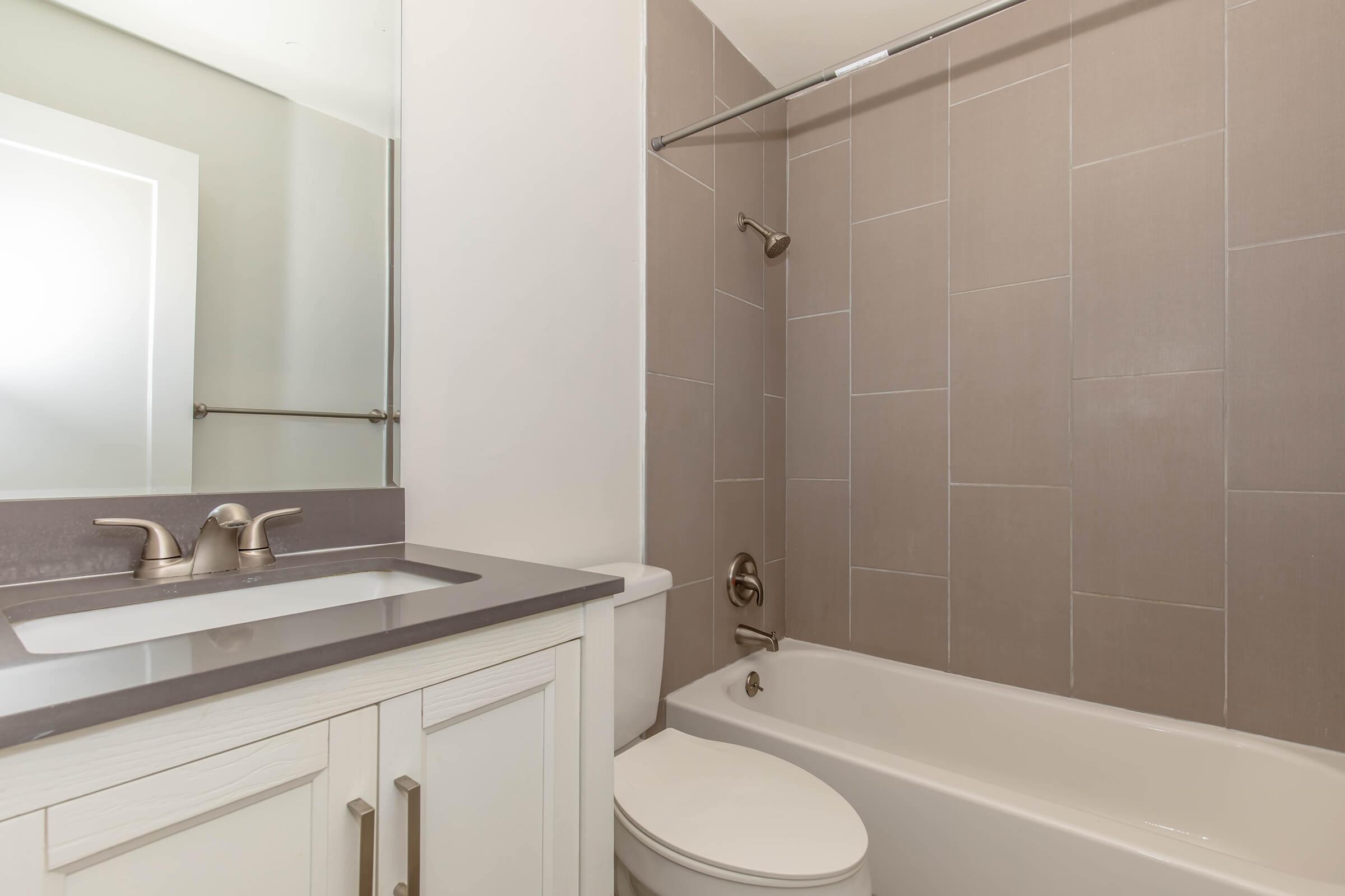 Modern bathroom featuring a clean design with a bathtub, a shower rod, and a large mirror above a sink with a faucet. The walls are tiled in a light gray color, and the fixtures have a contemporary style. Natural light enters from a window, enhancing the bright, airy feel of the space.