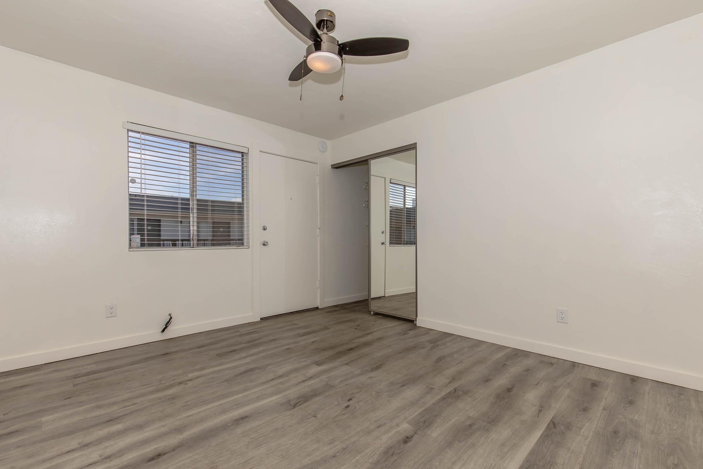 A sparsely furnished room with light-colored walls and wooden flooring. It features a ceiling fan, a window with horizontal blinds, and a mirrored closet door. Natural light fills the space, creating a bright and open atmosphere. The door is visible on one side, leading to the exterior.