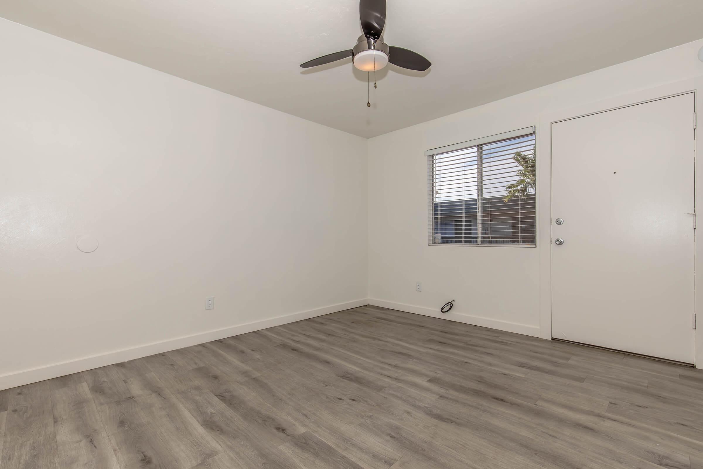 A minimalist interior of a room featuring light-colored walls, a ceiling fan, and wood-like flooring. A window with horizontal blinds allows natural light. The room includes a front door and a small wall outlet visible. The space is empty, showcasing potential for decorating and furnishing.