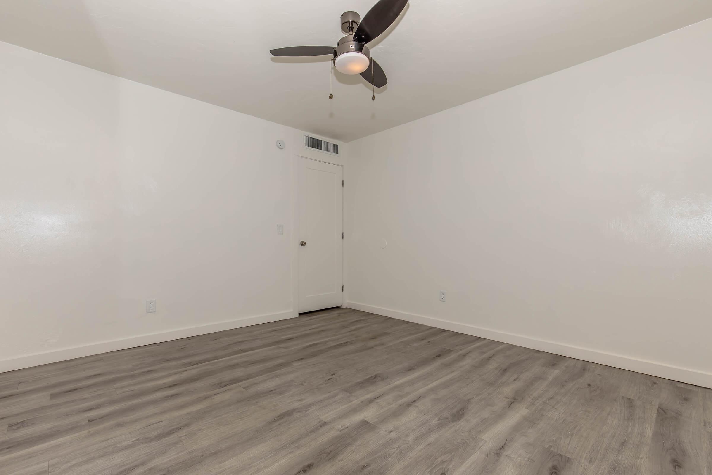 Empty room with light-colored walls and a wood-like laminate floor. A ceiling fan with three blades hangs in the center. A single closed door is visible on the right side, leading to another space. The room has no furniture or decorations, creating a minimalist and spacious appearance.