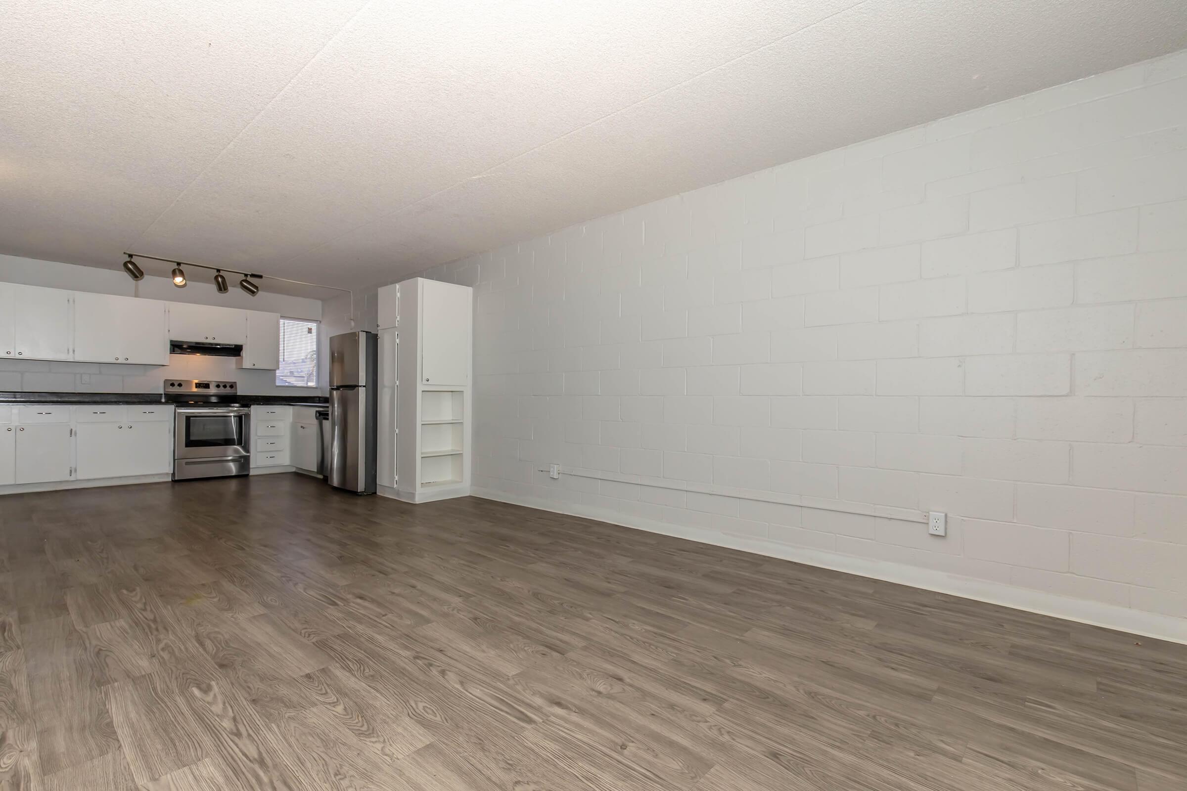A spacious, modern kitchen and living area with white walls and unoccupied wood-like flooring. The kitchen features stainless steel appliances, including a stove and refrigerator, along with white cabinetry. Natural light enters through a window, enhancing the open feel of the space.