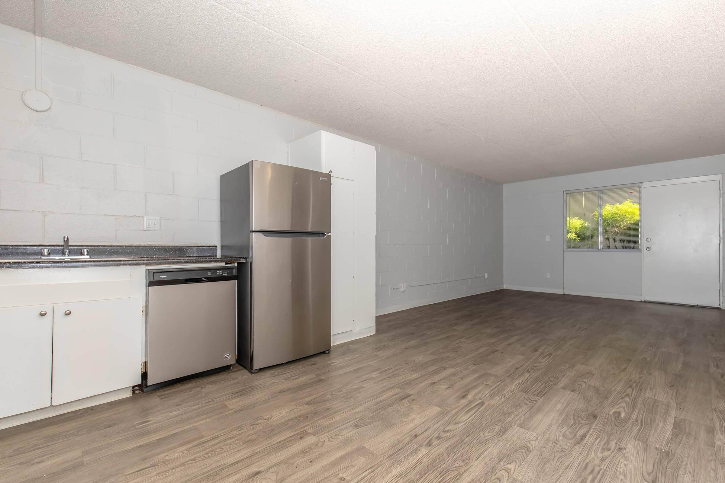 Interior view of a kitchen area featuring a stainless steel refrigerator and dishwasher, set against a light grey wall. The flooring is wood-like laminate. A window lets in natural light, while a door is visible at the far end, leading to another space. The atmosphere is simple and modern.