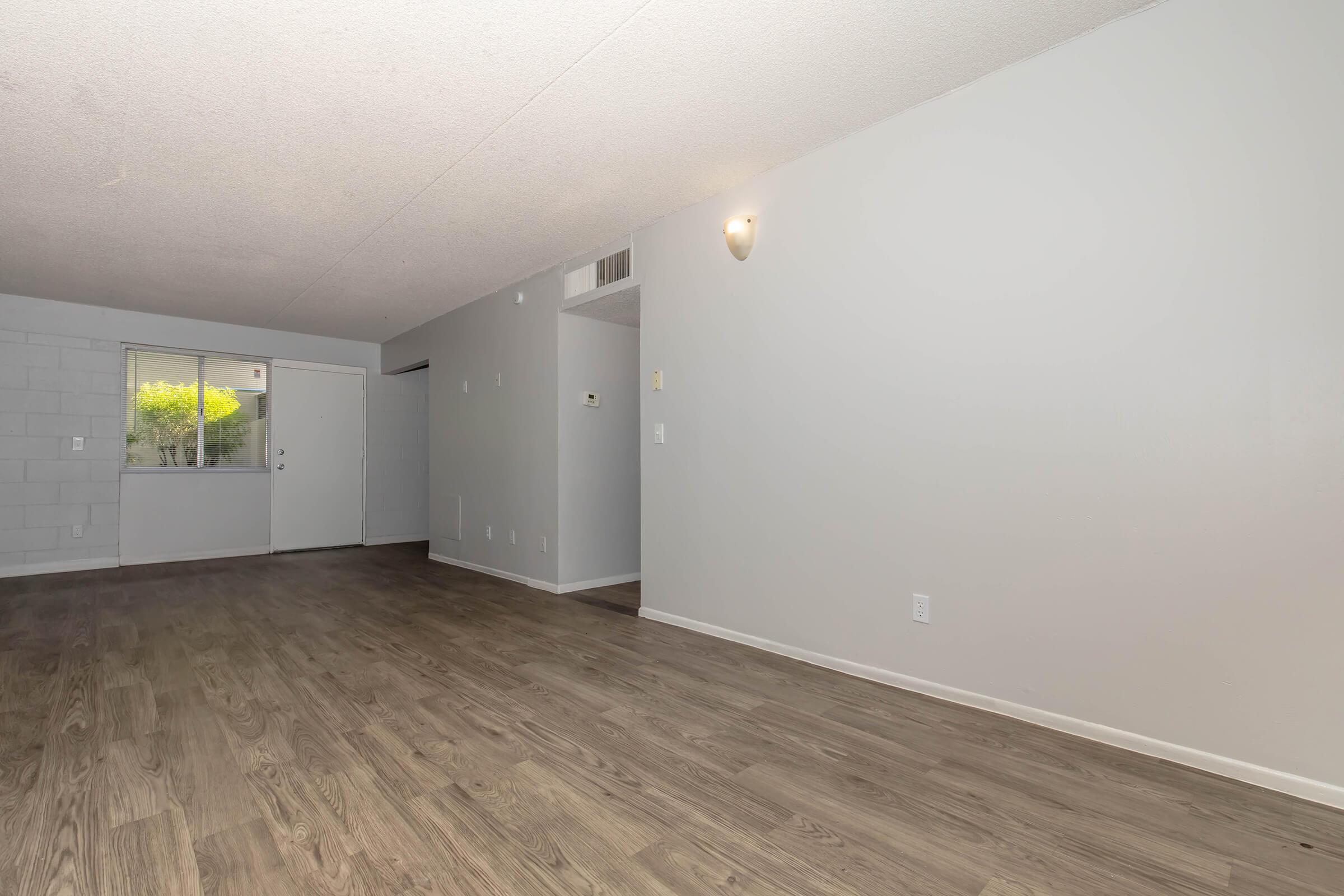 A spacious, empty living area with light gray walls and a wooden floor. A doorway on the left leads outside, and a small window on the left side lets in natural light. The room features a wall sconce for additional lighting, creating a bright, open feel.