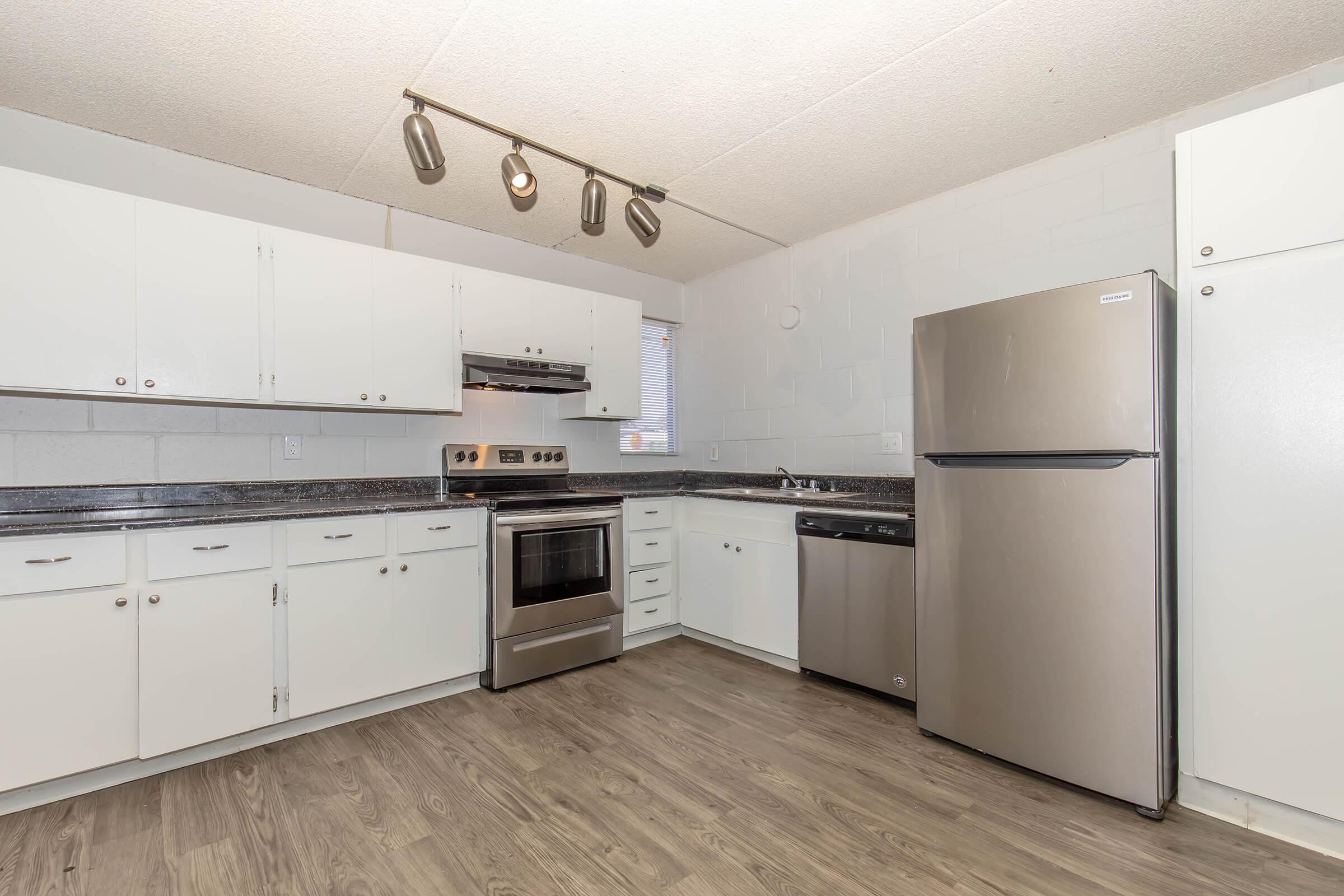 Modern kitchen featuring white cabinets, stainless steel appliances including an oven, refrigerator, and dishwasher, and a granite countertop with a sink. The space has track lighting and a large window, with laminate flooring for a clean, contemporary look.