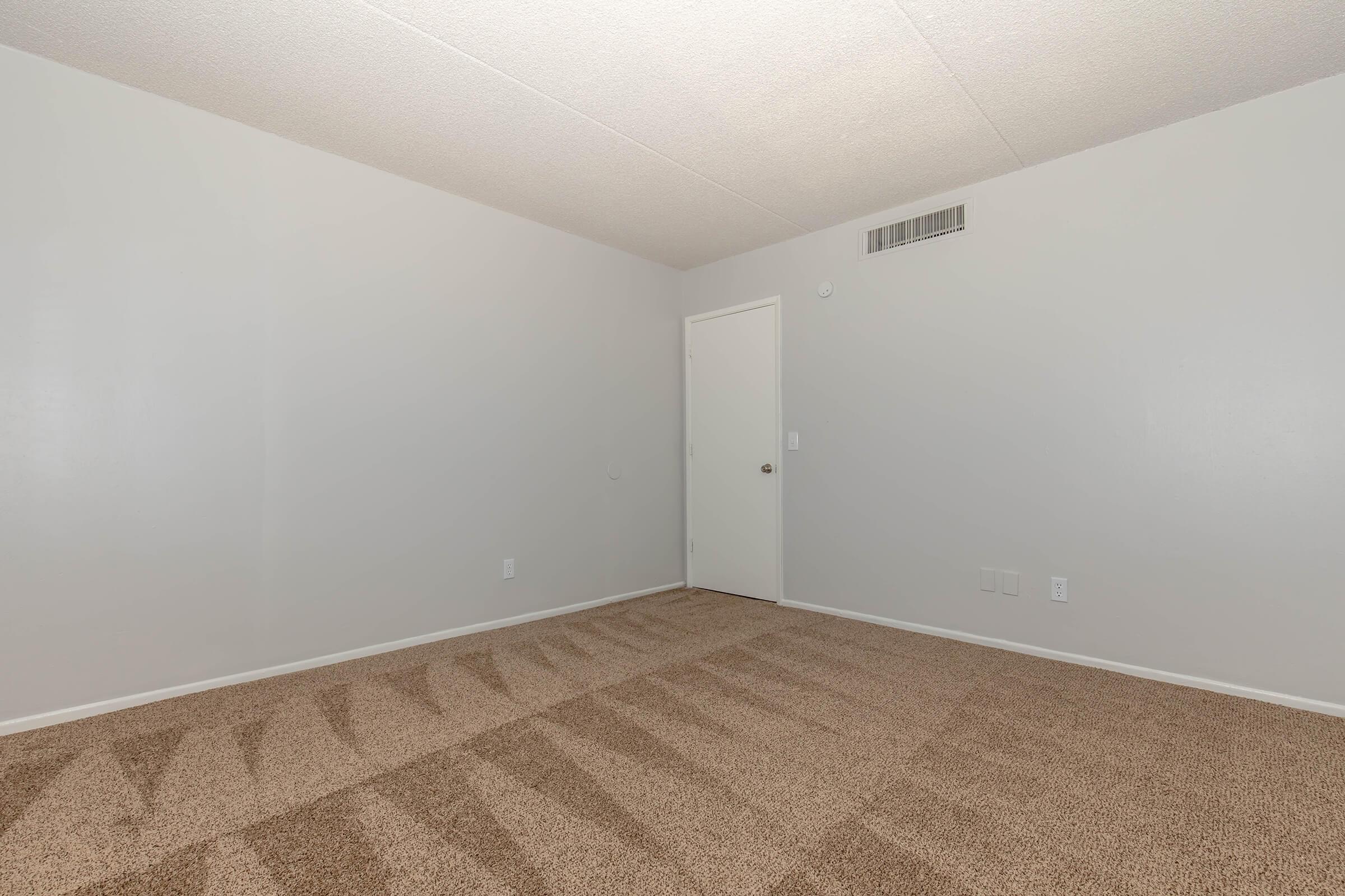 A vacant room with light gray walls and beige carpet. The space features a closed door on one wall and no furniture or decorations, creating a minimalist and spacious appearance. Natural light illuminates the area, highlighting the smooth ceiling and clean lines.