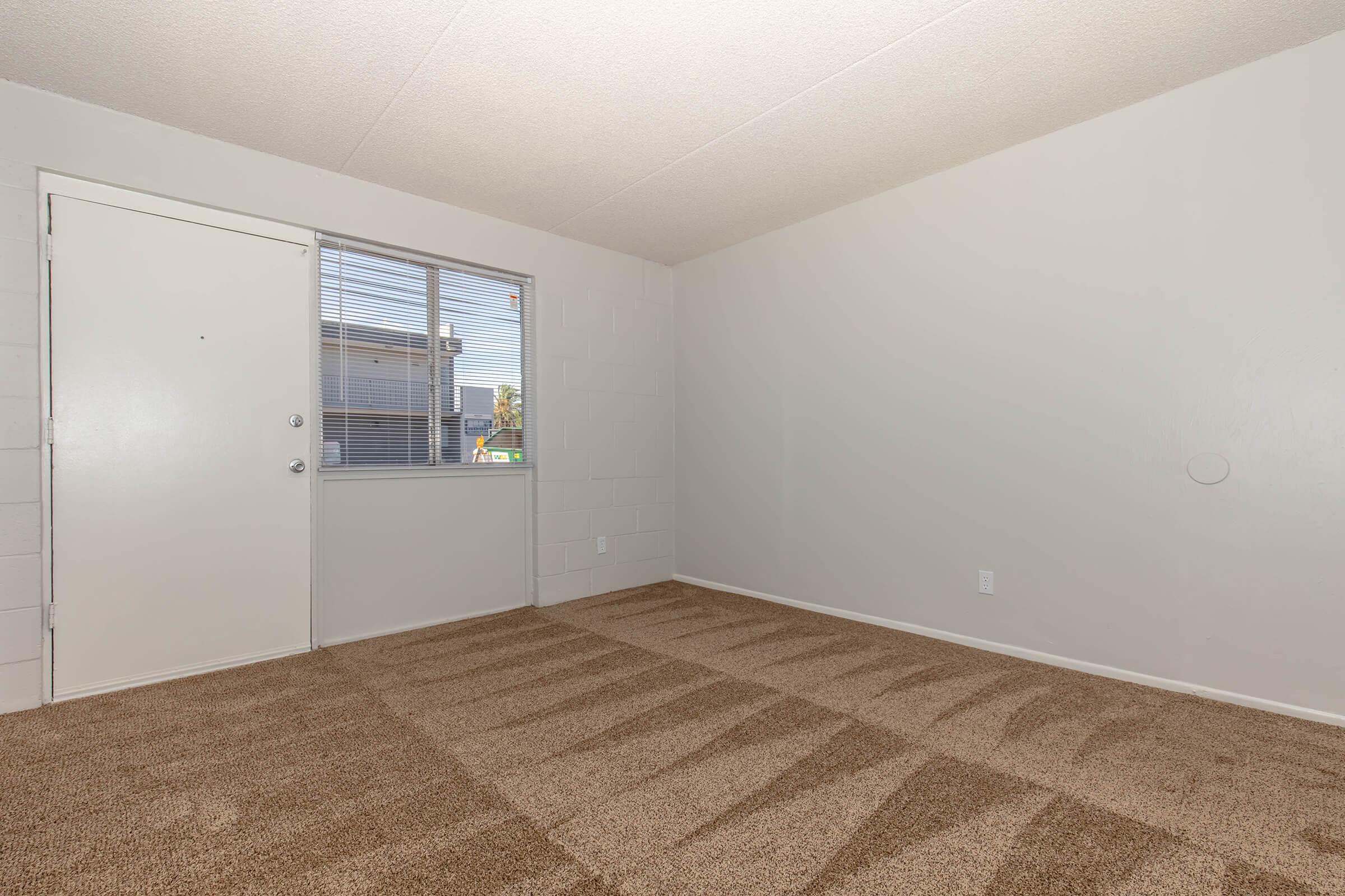 A clean, empty room with light beige carpet and light gray walls. The room features a window on the left side, allowing natural light to enter, and a plain door on the right. There are minimal furnishings or decor, creating a spacious and open feel.