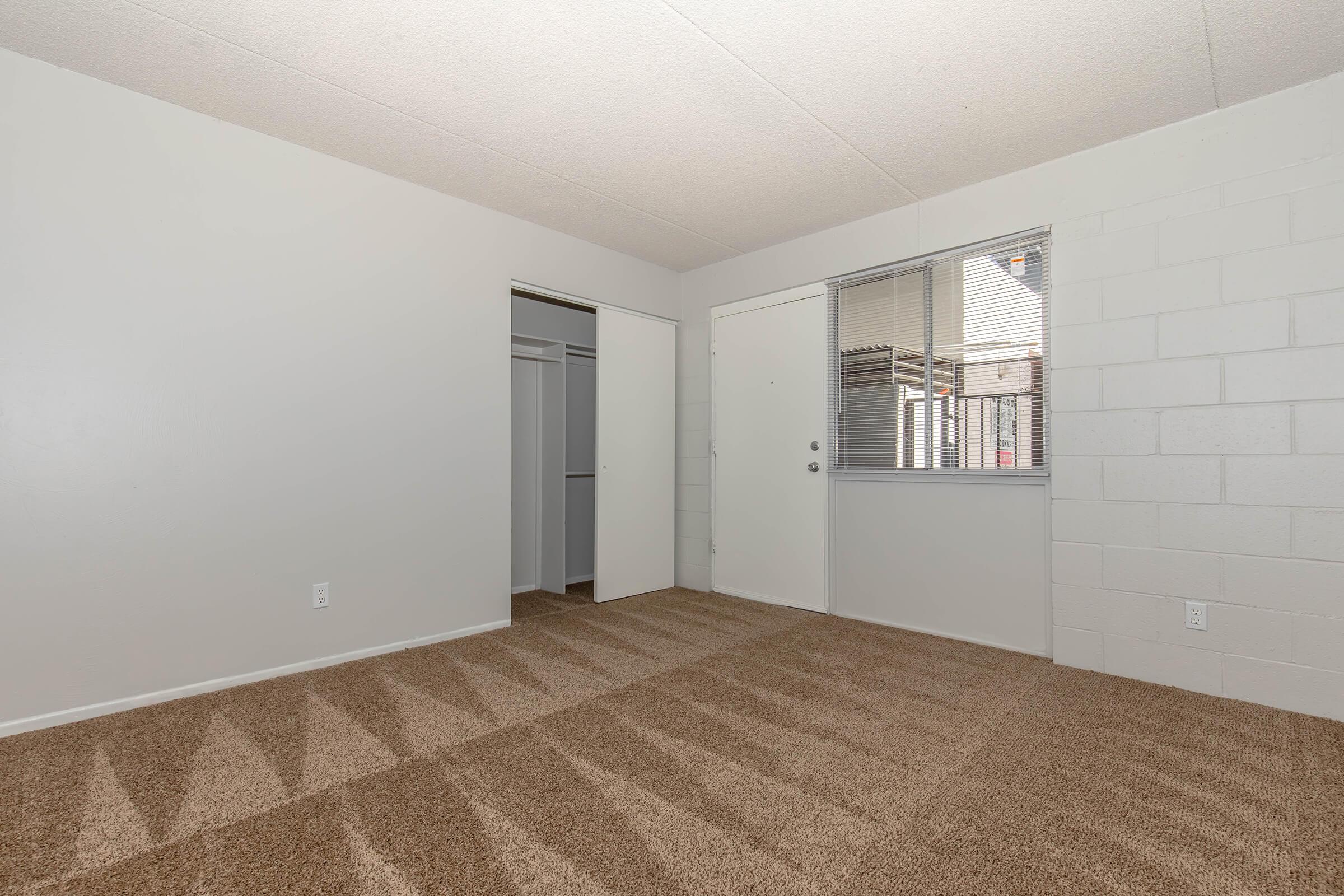 A spacious, empty room with light-colored walls and carpeted flooring. There's a closet with sliding doors on the left, and a window on the right letting in natural light. A door leads to the outside, creating a bright and open feel. The room is ready for furnishing.