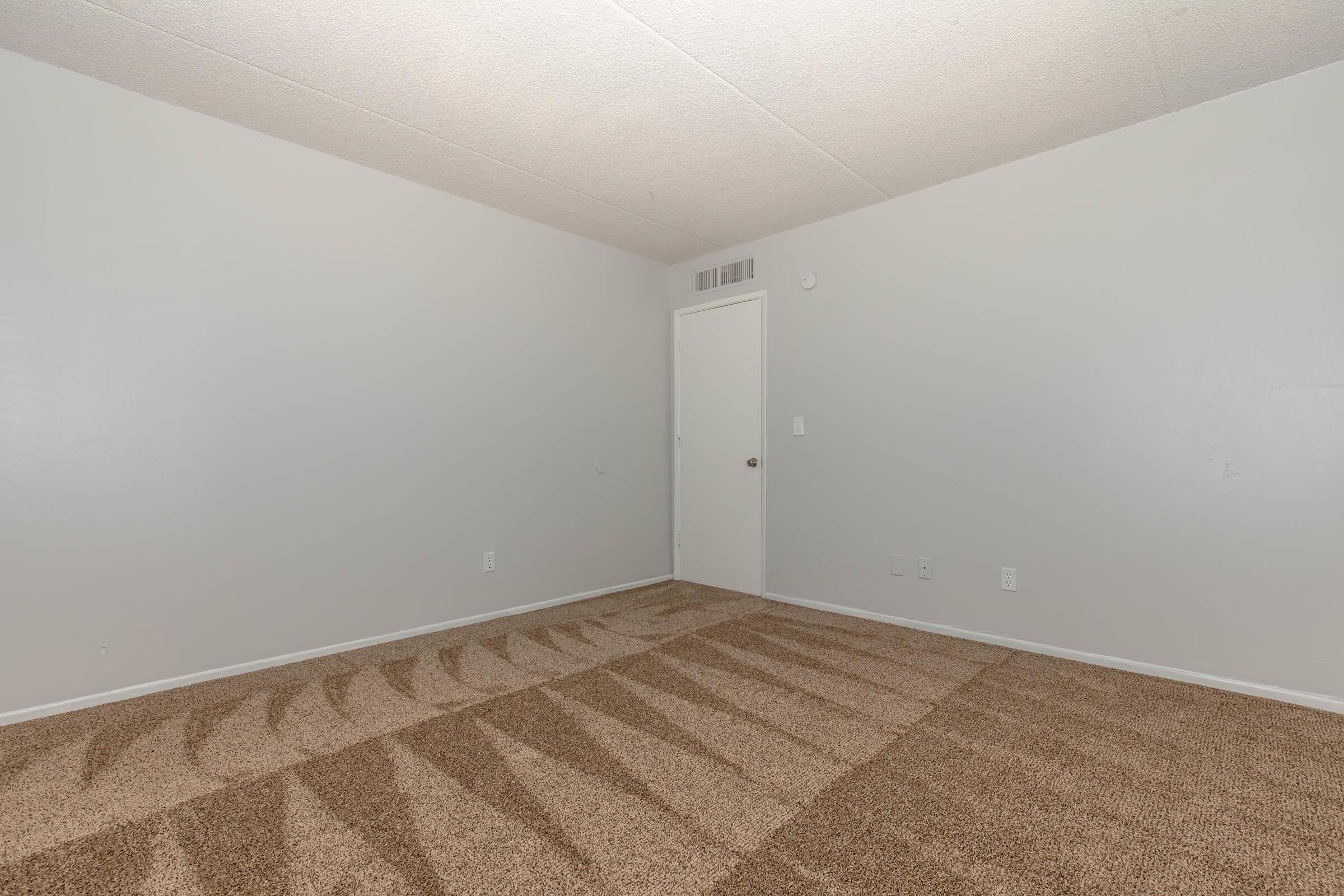 Empty room with light gray walls and a tan carpet. A single white door is visible on the far wall, and there are no furnishings or decorations in the space. The floor shows distinct shadows from light sources, emphasizing the room's emptiness.