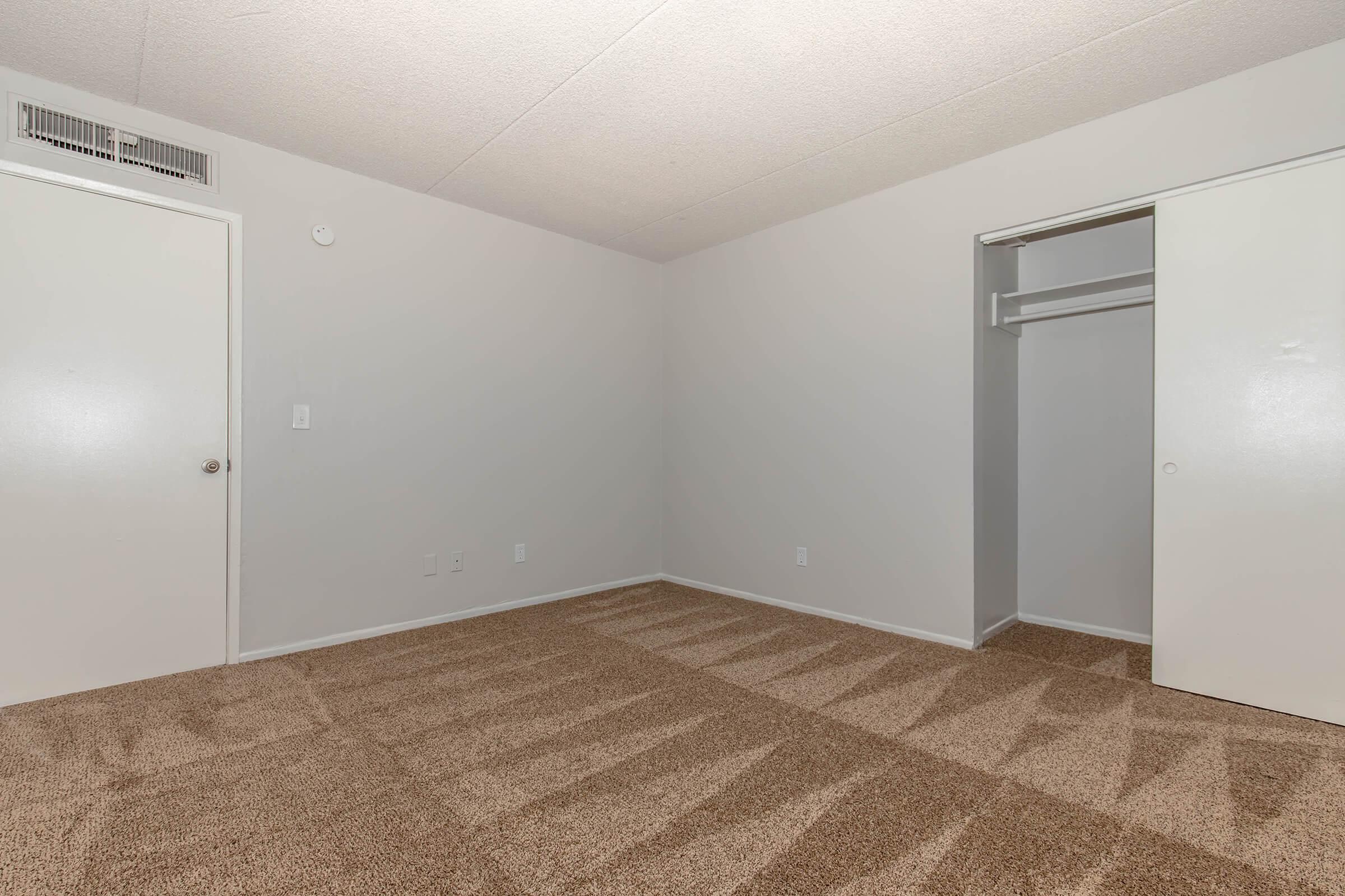 A vacant room with light gray walls and beige carpet. There is a white door leading outside and a closet with sliding doors on the right side. The room is well-lit and spacious, with no furniture present. The flooring shows shadows from the room's structure.