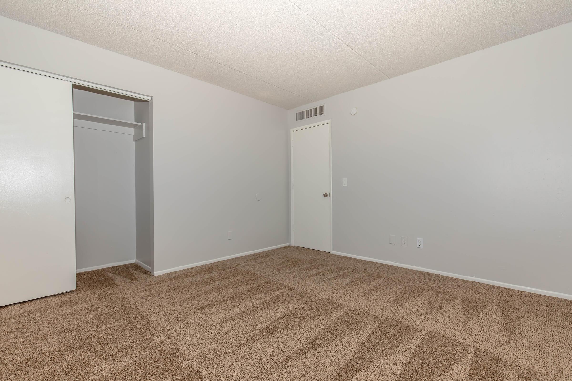 Empty room with light gray walls and a tan carpet. One side features a closet with sliding doors, while the opposite side has a white interior door. The ceiling has a textured finish, and there are no furnishings or decorations in the space, giving it a spacious and minimalistic appearance.