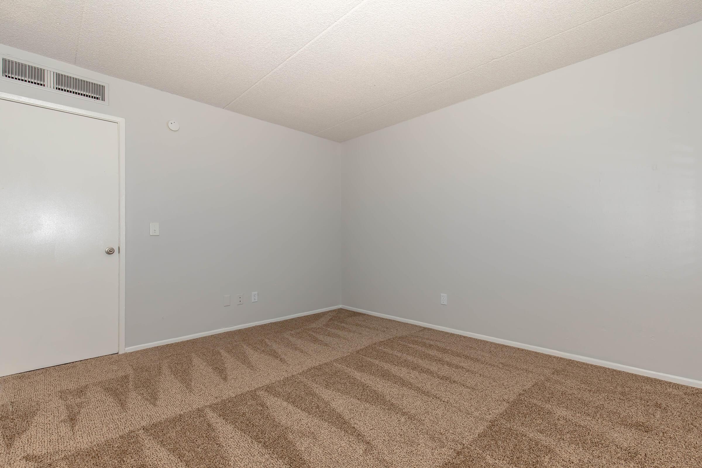 A vacant room with light gray walls and a plush beige carpet. The room features a closed white door and a small air vent on the ceiling. Sunlight creates subtle shadows on the carpet, adding depth to the space. The overall ambiance is neutral and minimalist, providing a blank canvas for decoration.
