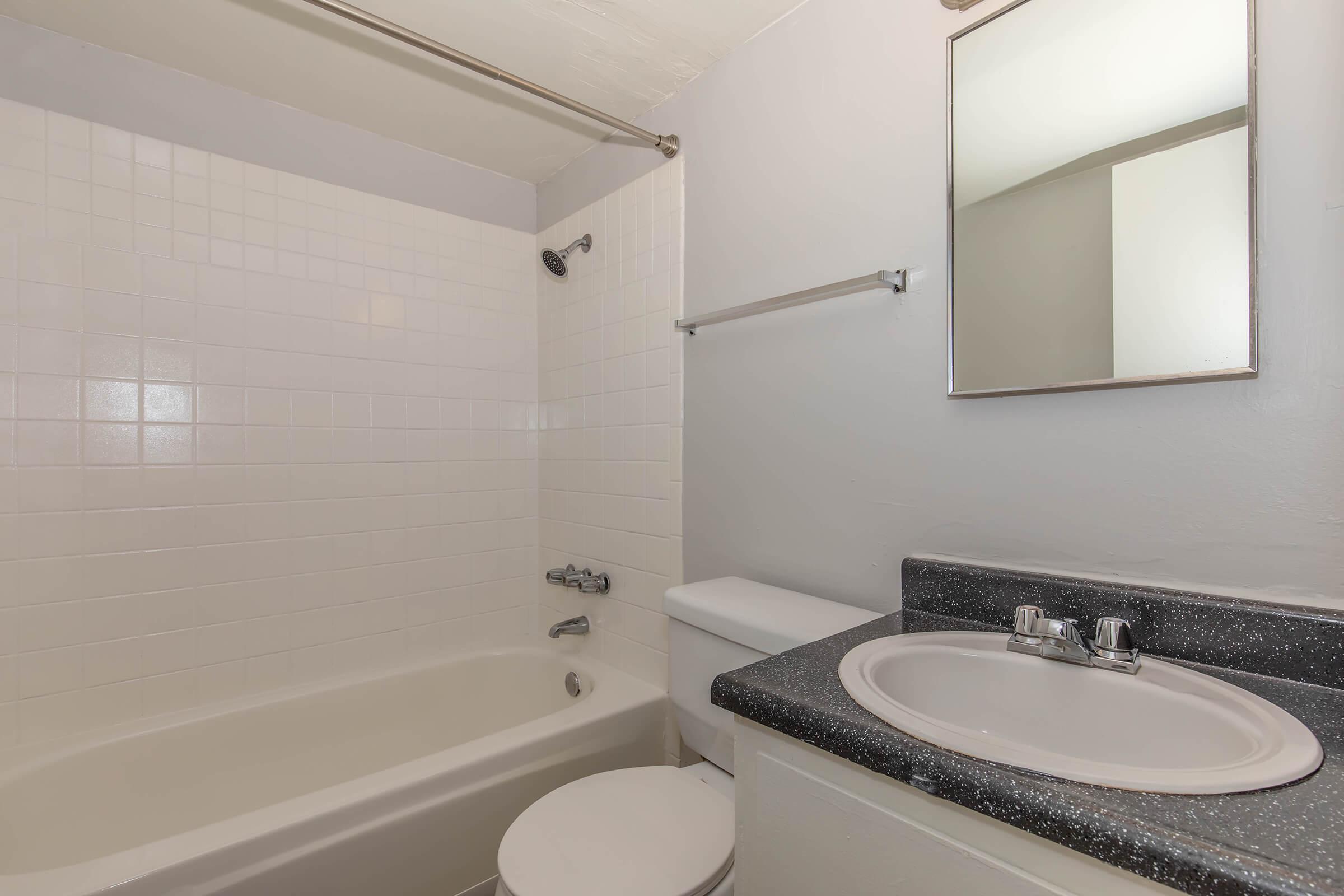 A clean and simple bathroom featuring a white tiled shower and bathtub, a white toilet, and a circular mirror above a dark speckled countertop with a sink. The walls are painted a light color, and there is a towel bar on the wall. The overall design is minimalist and functional.
