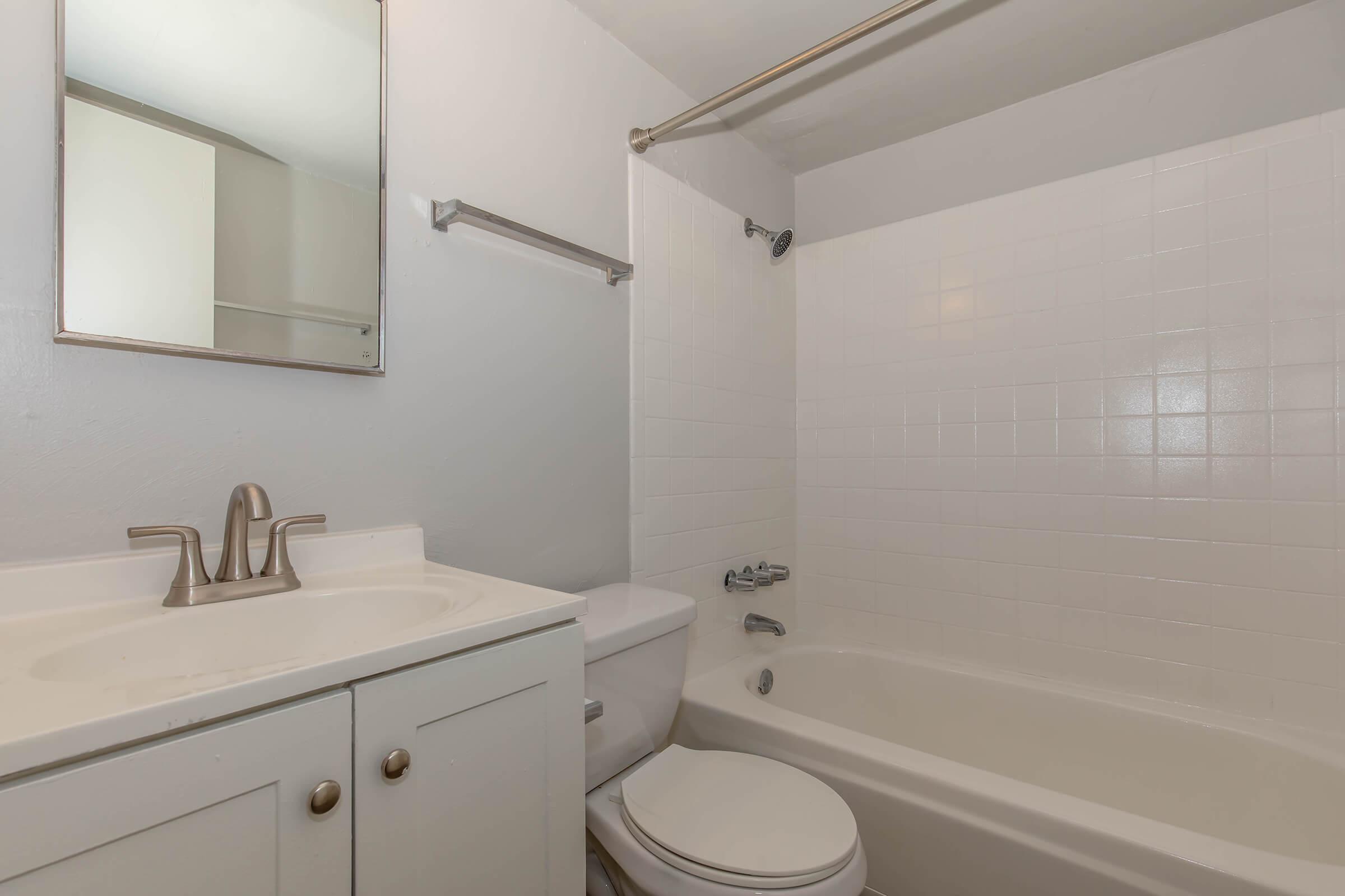 A clean and simple bathroom featuring a bathtub, toilet, and a sink with a mirror above it. The walls are tiled white, and there’s a shower rod installed above the tub. The cabinetry is in a light color, contributing to the overall bright and airy feel of the space.