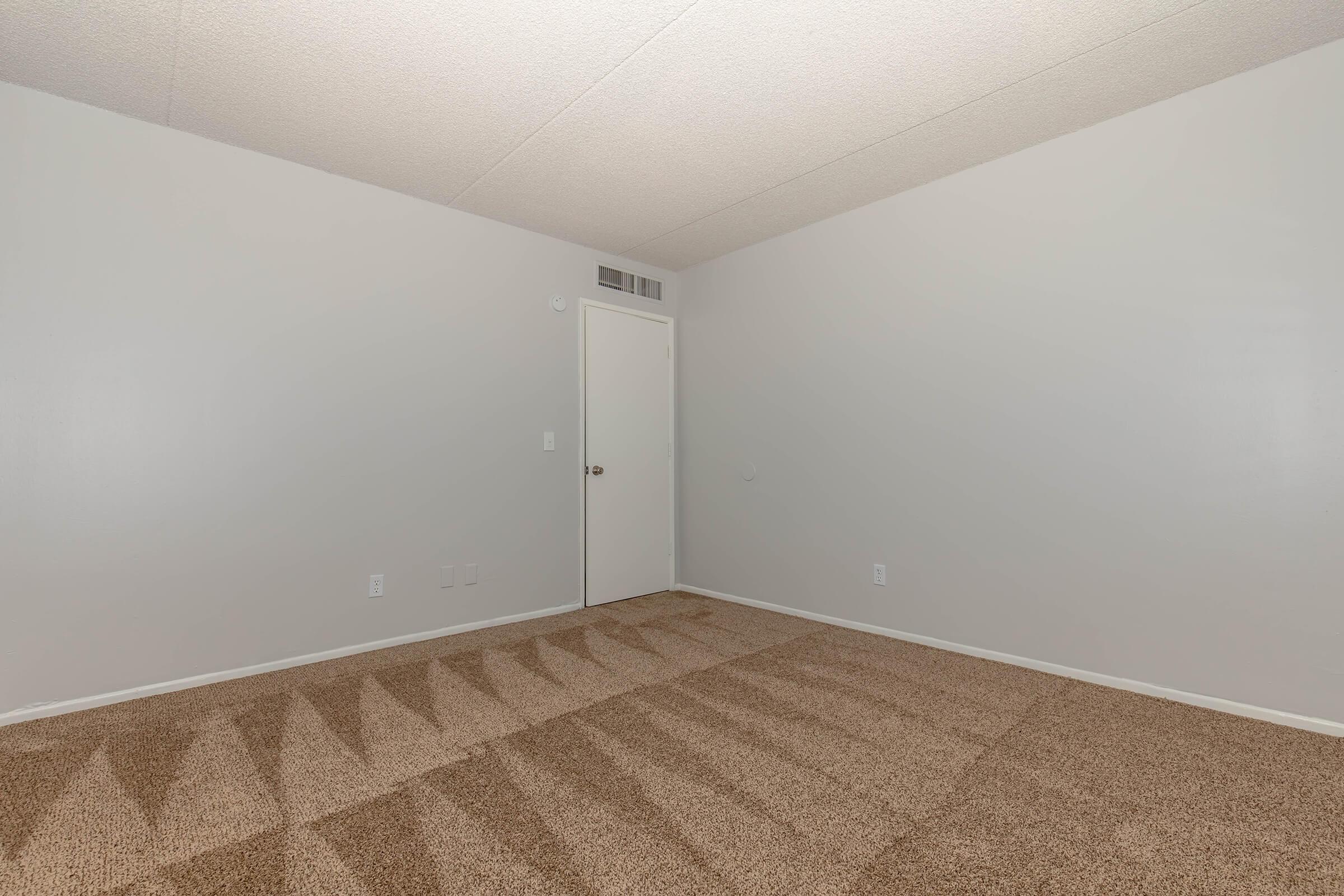 Empty room with light gray walls and a beige carpeted floor. A white door is visible on one wall, and there are no furnishings or decorations. The ceiling has a textured finish, and the overall ambiance is clean and spacious, with natural light creating subtle shadows on the carpet.