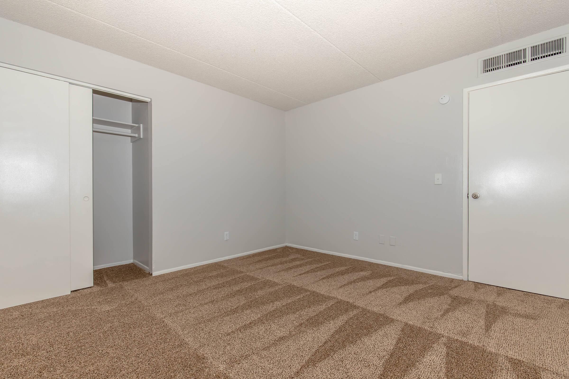 A spacious, empty bedroom with light gray walls and beige carpet. A closed door leads to a closet on the left, while another door is visible on the right. The room is well-lit with no furniture, providing a blank canvas for decoration.