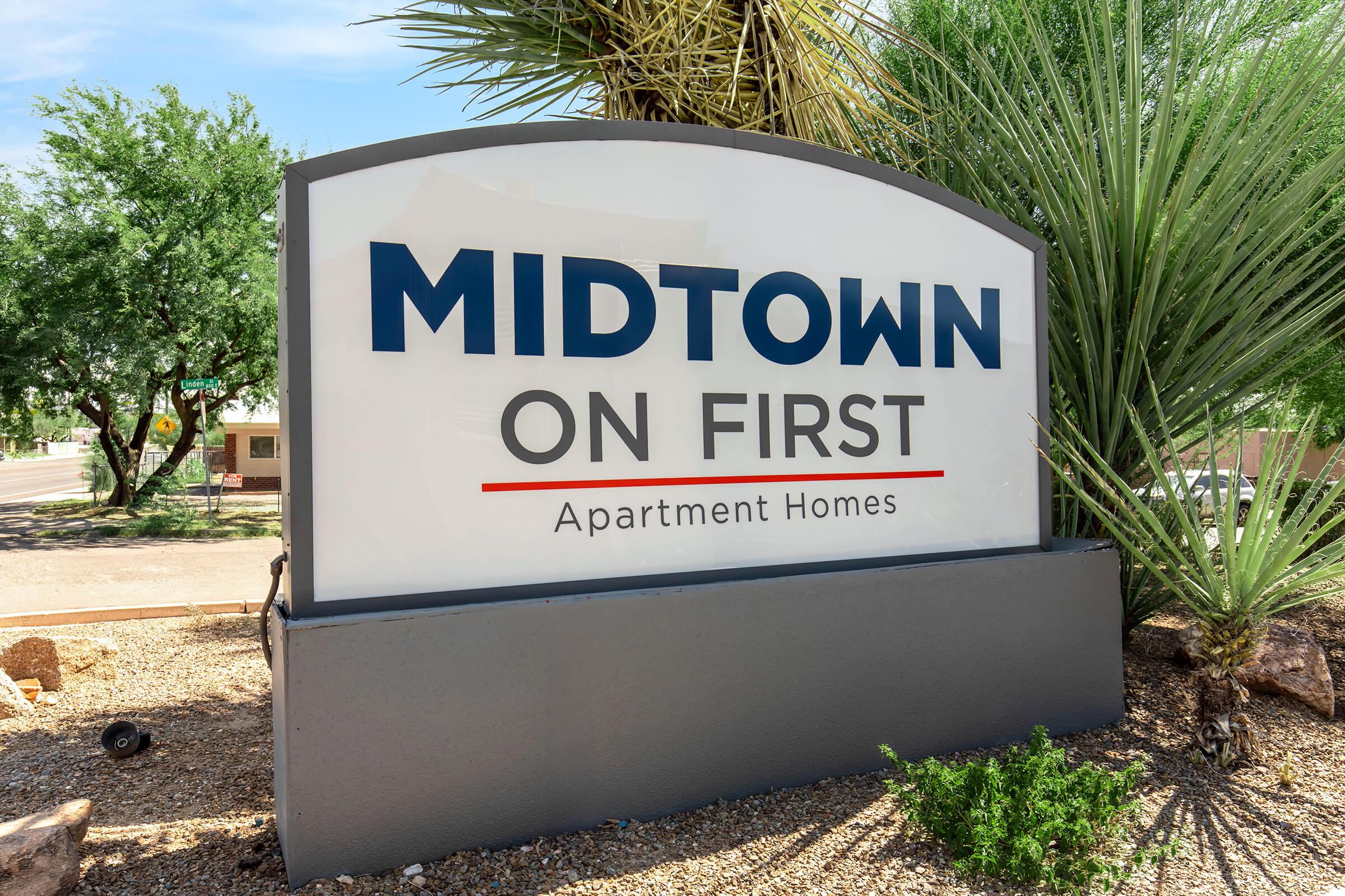 Sign for "Midtown on First" Apartment Homes, featuring bold blue and red lettering on a white background, surrounded by desert landscaping with palm trees and shrubs. The sign is positioned in a well-lit outdoor area, indicating a residential complex.