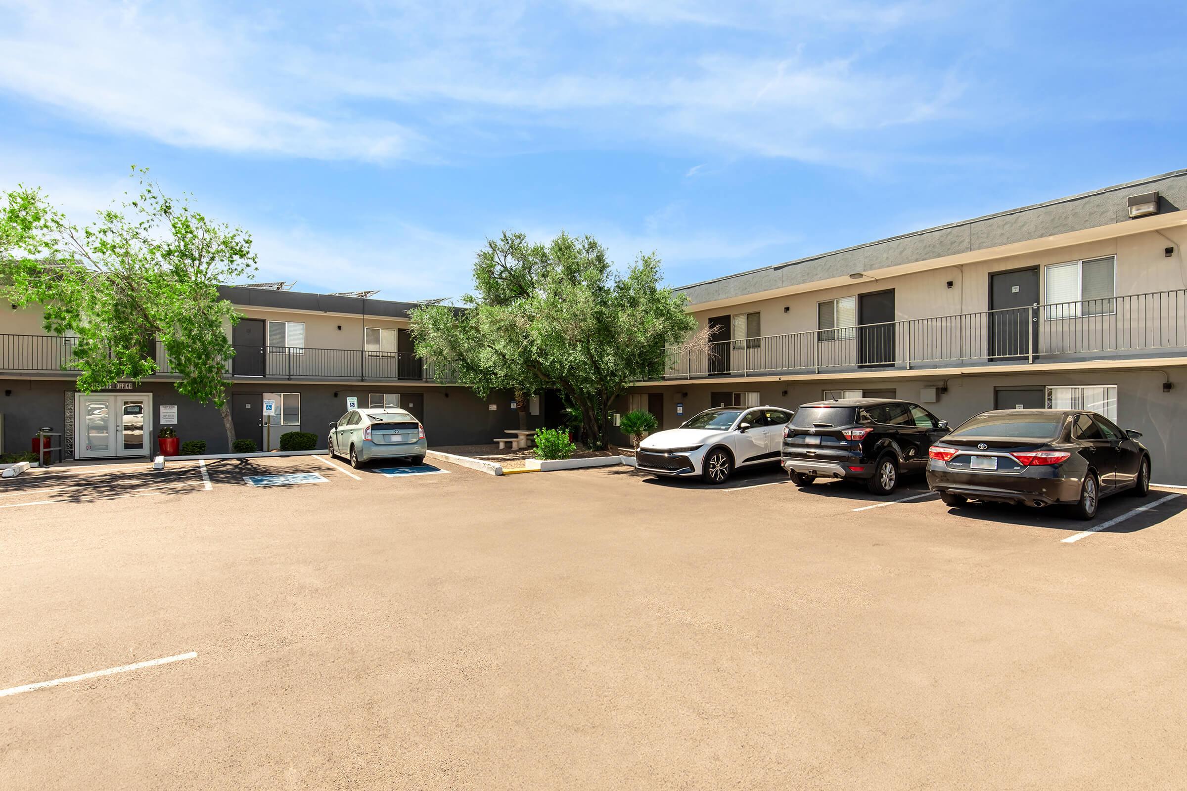 A parking lot in front of a two-story apartment building. The building features multiple balconies and a light gray exterior. Several cars are parked in the lot, including a white electric vehicle, a black SUV, and a silver sedan. Green trees are visible in the area, under a clear blue sky.