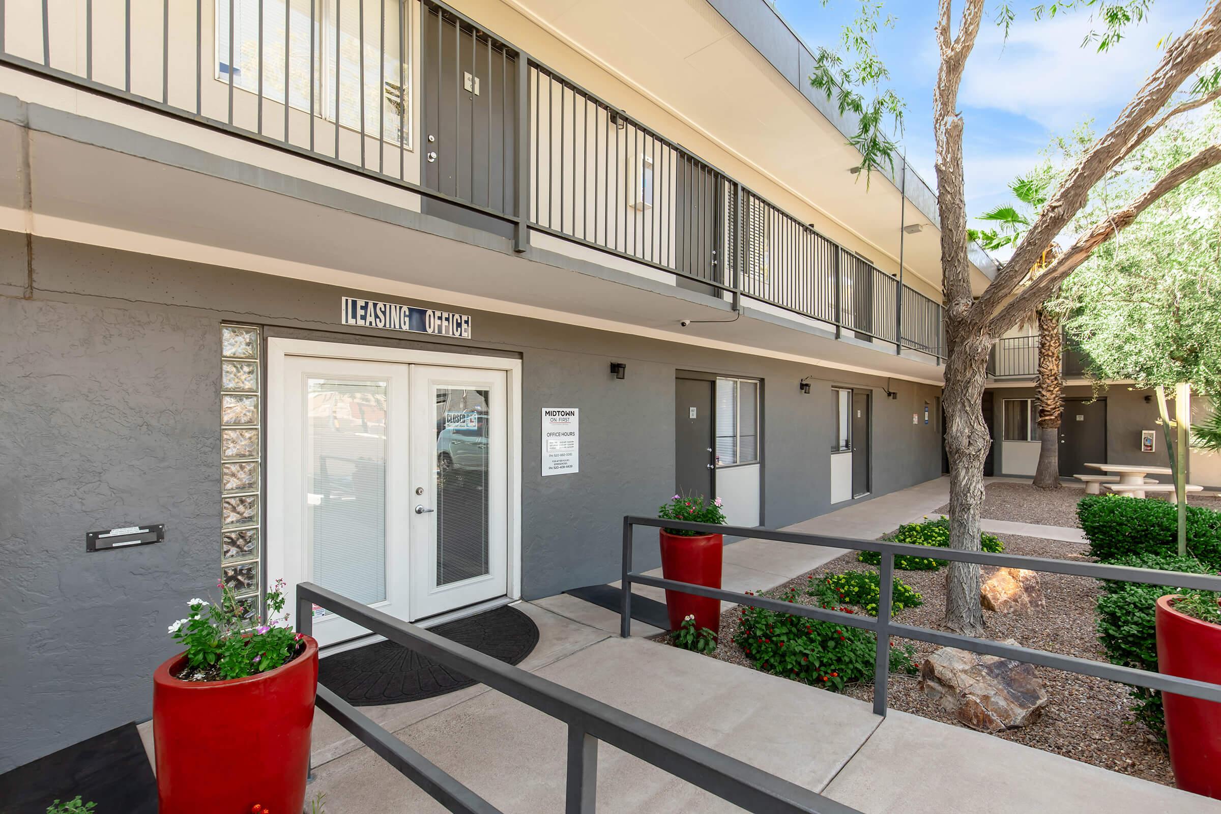 Exterior view of a low-rise building with a leasing office entrance. The office has glass doors and is flanked by two red planter pots with greenery. The building features balconies, light gray walls, and is set in a landscaped area with pathways and small trees. Sunlight brightens the scene.
