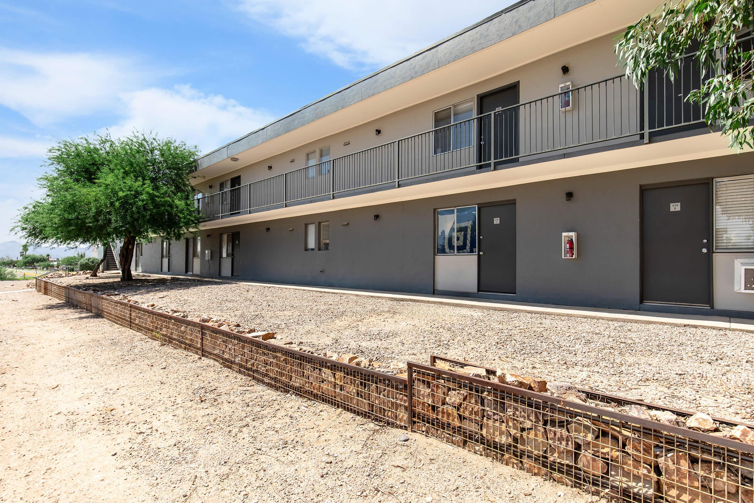 A low-rise apartment building with multiple units. Each unit has a door and small windows. The exterior is painted in a neutral color, and there is a landscaped area with gravel and a tree in front. The sky is clear with some clouds, suggesting a bright day.