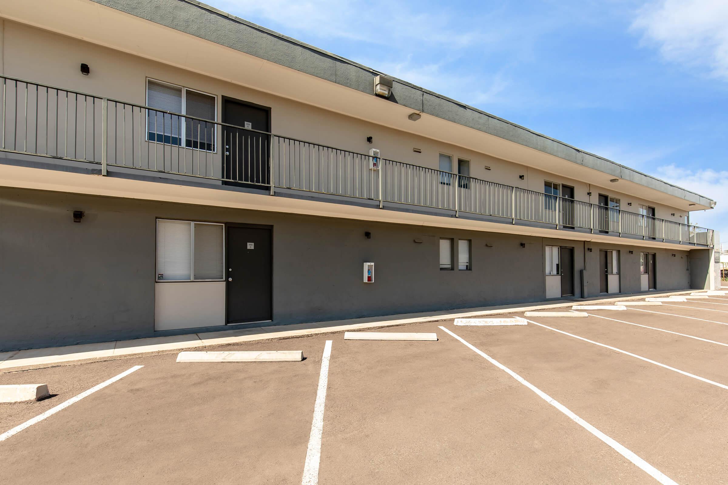 A low-rise motel building with a gray facade and multiple rooms. Each room features a balcony with metal railings. The parking lot in front has several empty parking spaces, and blue sky with a few clouds is visible in the background.