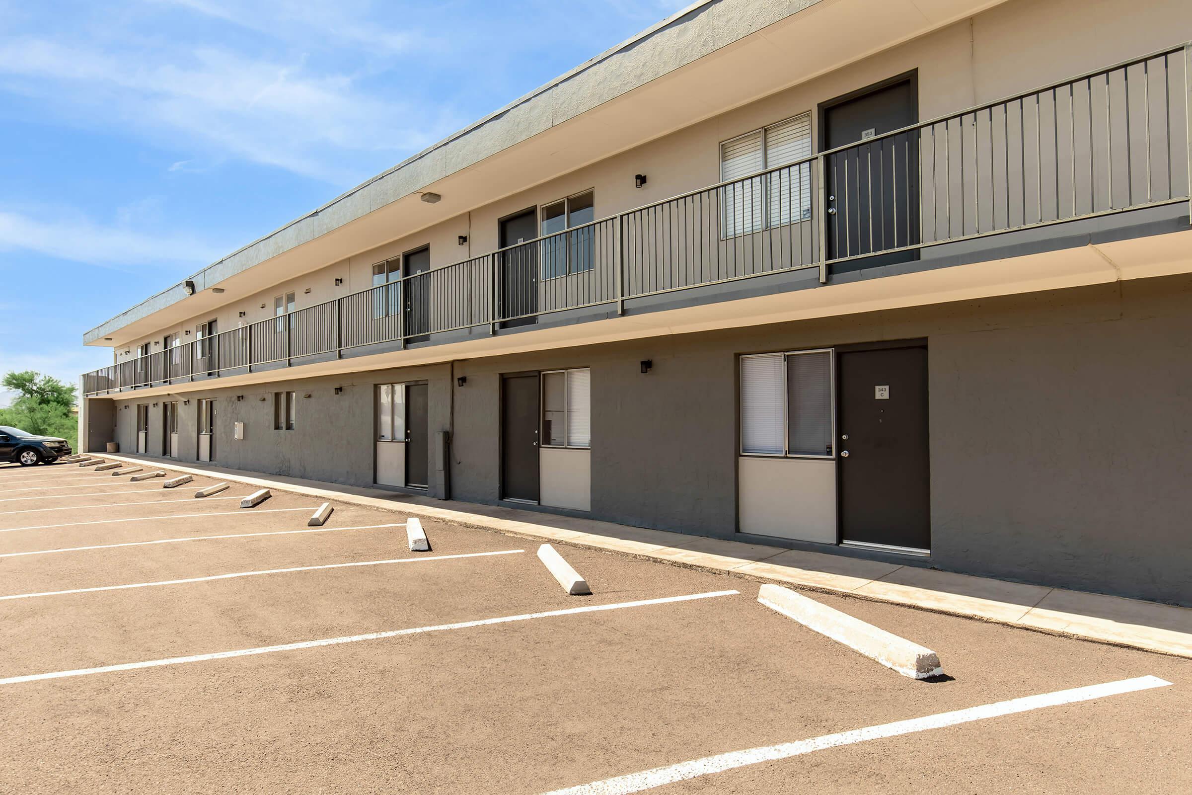 A modern two-story motel building with several doors and balconies, located next to a parking lot with white parking spaces. The exterior features a mix of gray and beige colors, and clear blue skies are visible overhead.