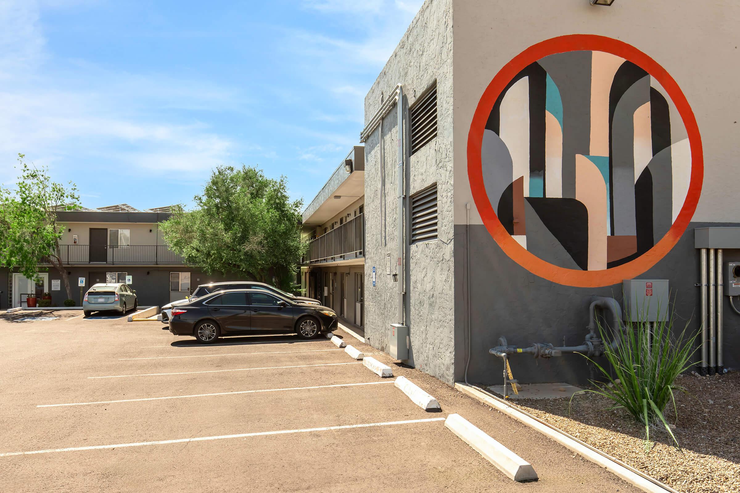 View of a parking lot with several parked cars and a modern building featuring a colorful mural. The mural consists of abstract shapes in shades of blue, gray, and orange, set against a textured wall. Lush greenery is visible in the background, under a clear blue sky.