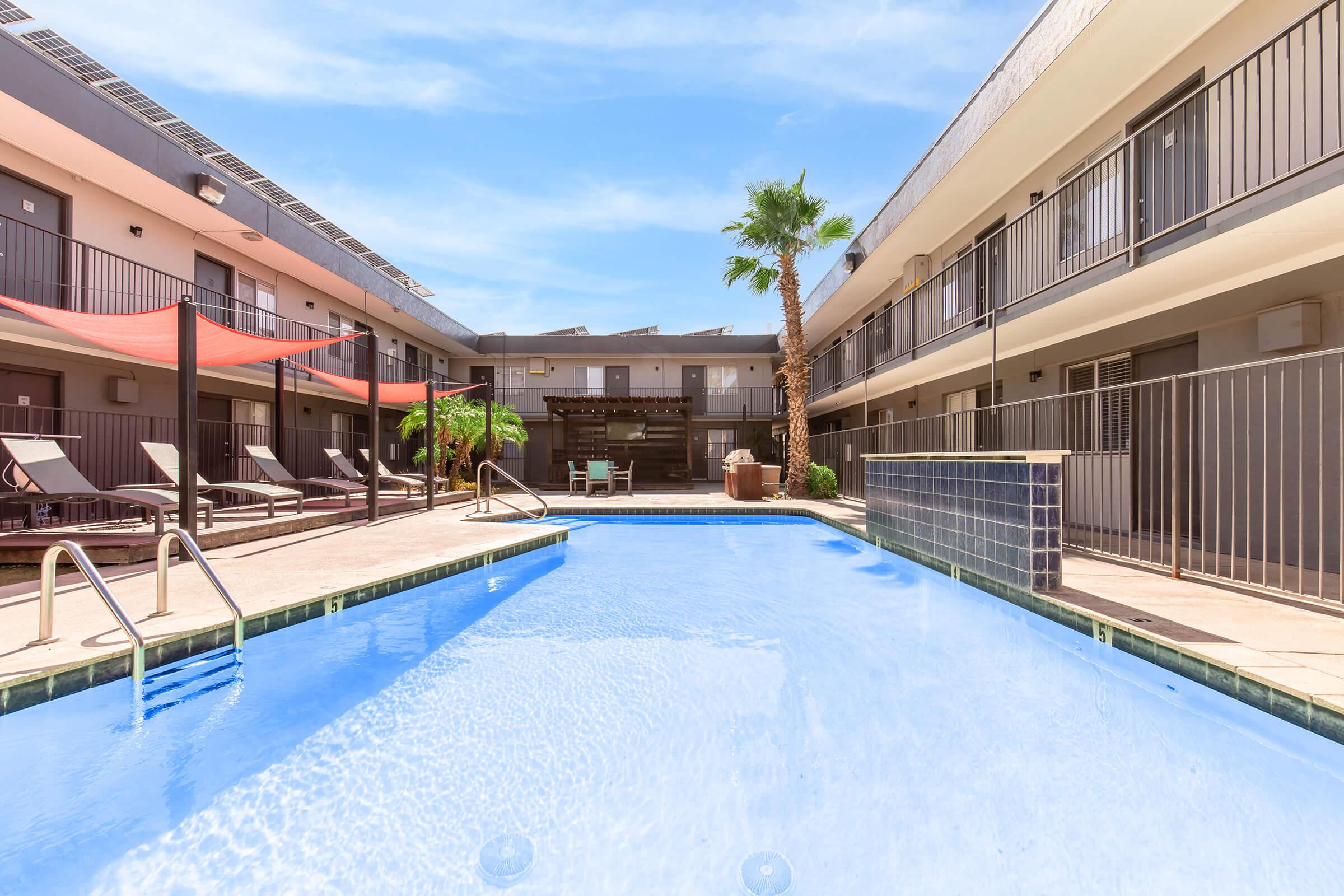 A clean, inviting swimming pool surrounded by lounge chairs and tropical plants, situated in an outdoor area of a modern hotel. The pool reflects blue skies, and there are palm trees and shaded seating areas nearby, creating a relaxing atmosphere in the courtyard.